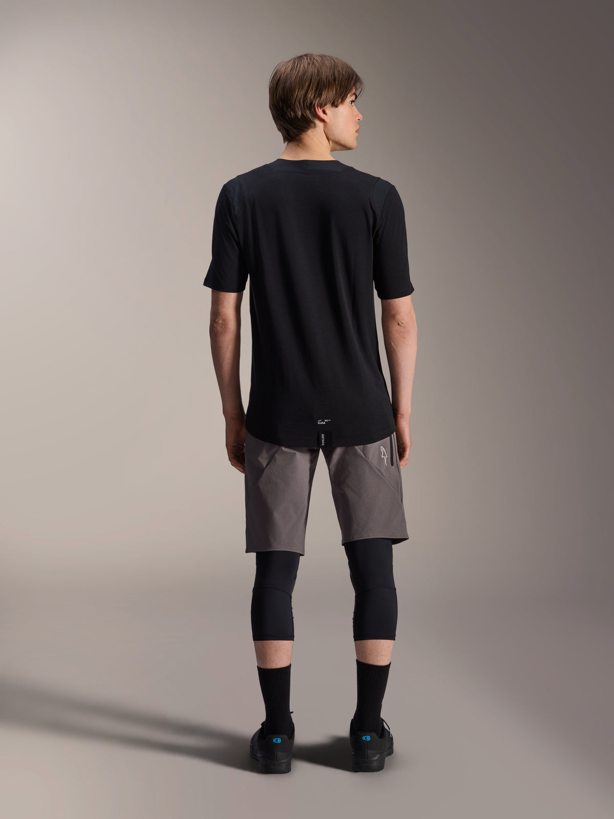 Alpinestars A-Dura Dri Wool kortærmet jersey, sort, set bagfra på model med grå MTB-shorts og sorte knæbeskyttere, med en længere drop tail og atletisk pasform til off-road cykling