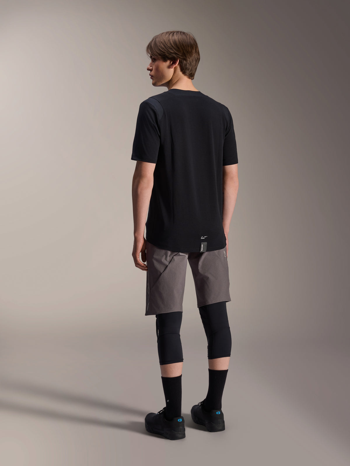 Alpinestars A-Dura Dri Wool Kortærmet Jersey, sort, set bagfra på model, båret med grå mountainbike-shorts og sorte knæbeskyttere, med forlænget rygstykke og diskret logo, teknisk MTB-beklædning