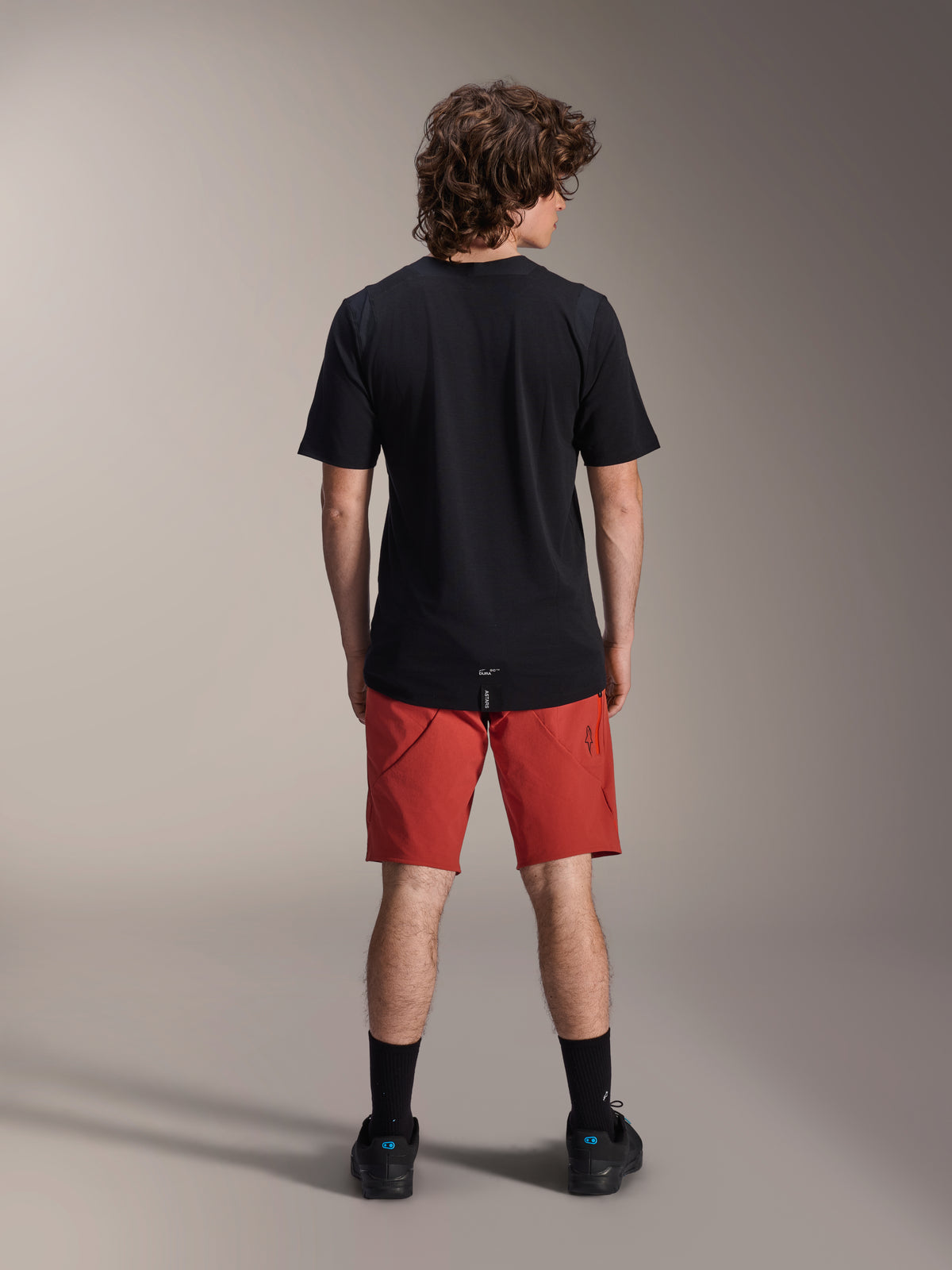 Male model wearing Alpinestars A-Dura Elite Shorts in Dark Orange and a black A-Dura Dri Wool Short Sleeve Jersey, seen from the back, paired with black socks and cycling shoes, professional mountain bike apparel
