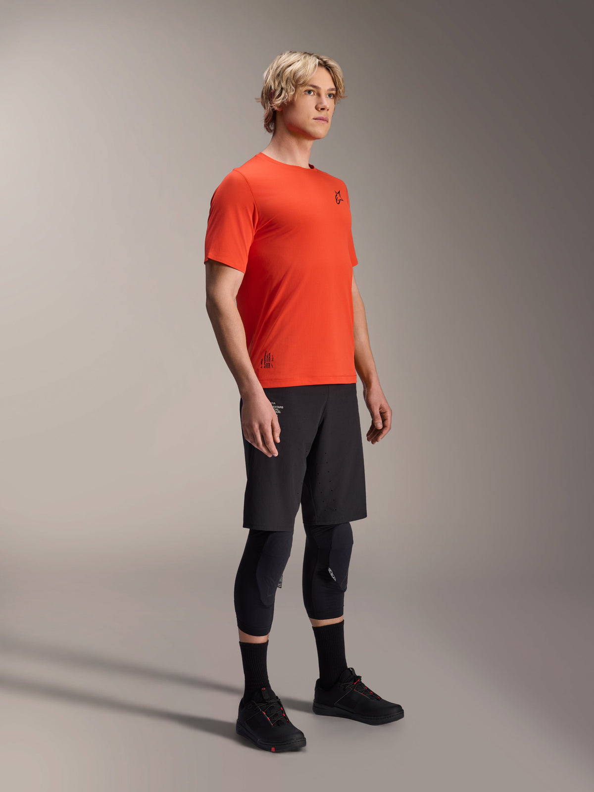 Alpinestars A-Aria Astar kortærmet trøje i Spicy Orange, båret af en model med sorte MTB-shorts og knæbeskyttelse, letvægts åndbart mountainbike-tøj, levende orange med minimal sort branding