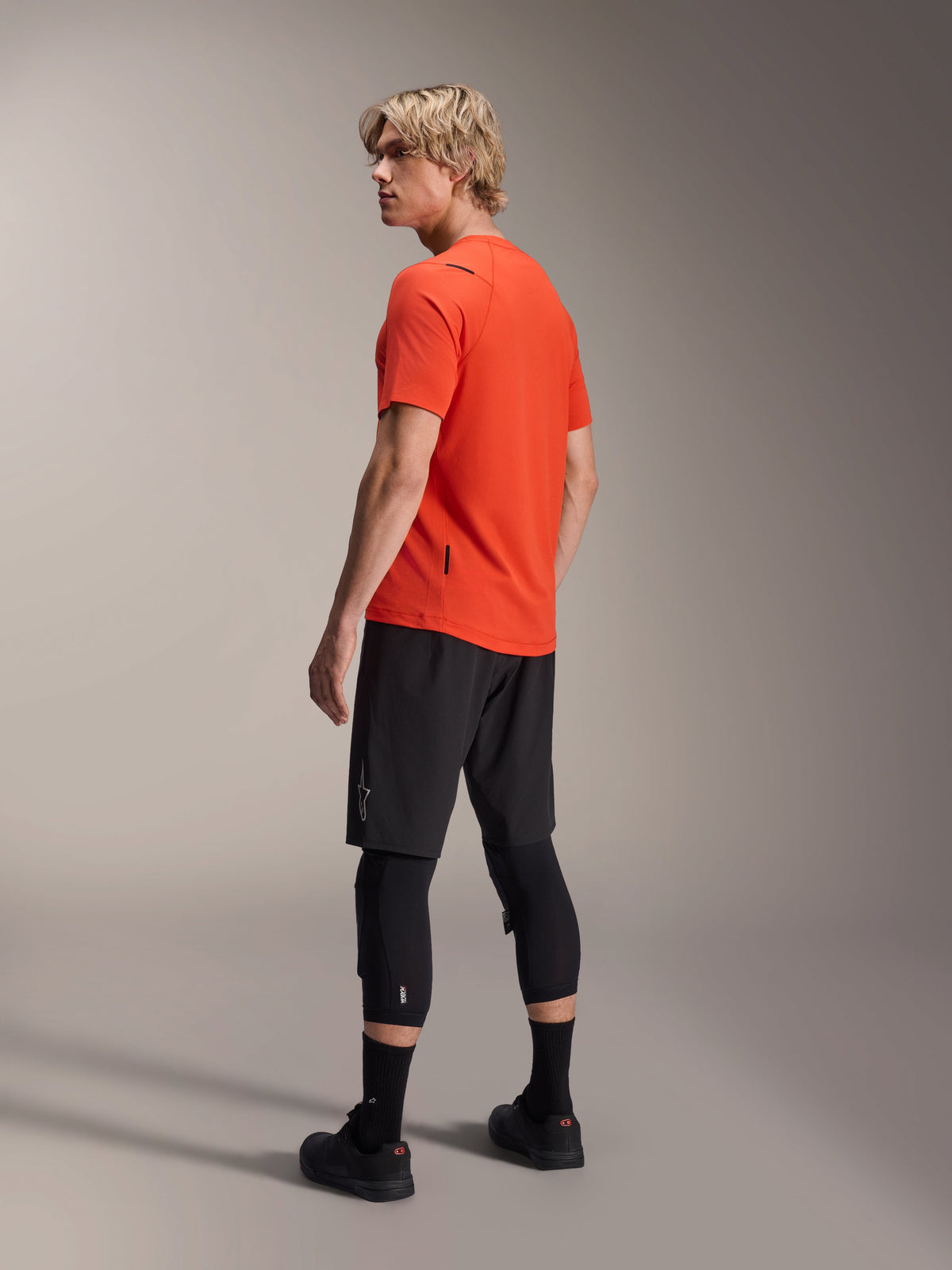 Alpinestars A-Aria Astar kortærmet trøje, Spicy Orange, trefjerdedelsvisning bagfra af model der kigger over skulderen, kombineret med sorte MTB-shorts og knæbeskyttere, performance mountainbike-tøj