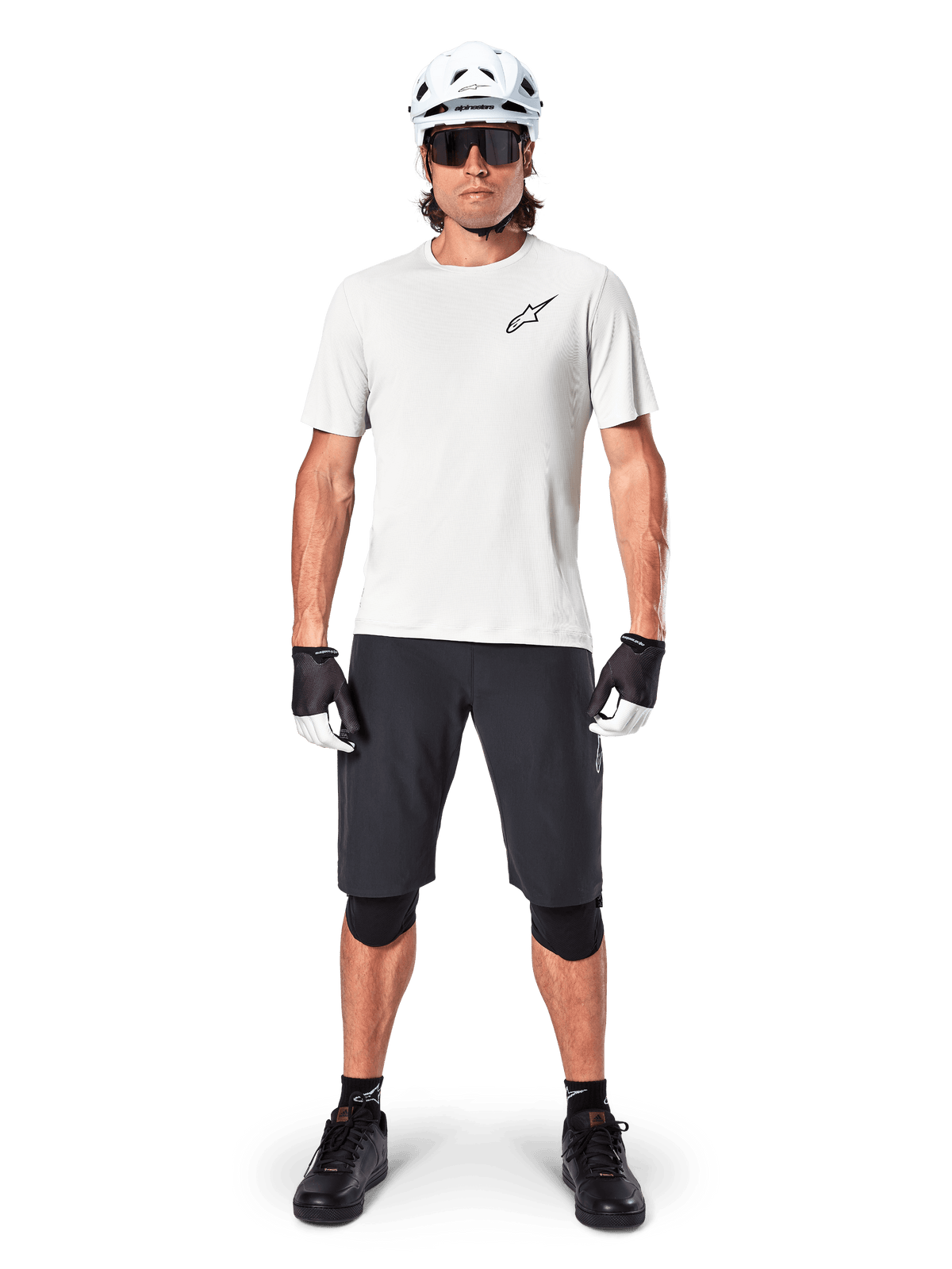 Male cyclist wearing Alpinestars A-Aria Astar Short Sleeve Jersey in White Dove, featuring a black Astars logo on the chest, paired with black mountain bike shorts, white helmet, sunglasses, black knee guards, and gloves, complete off-road cycling outfit.