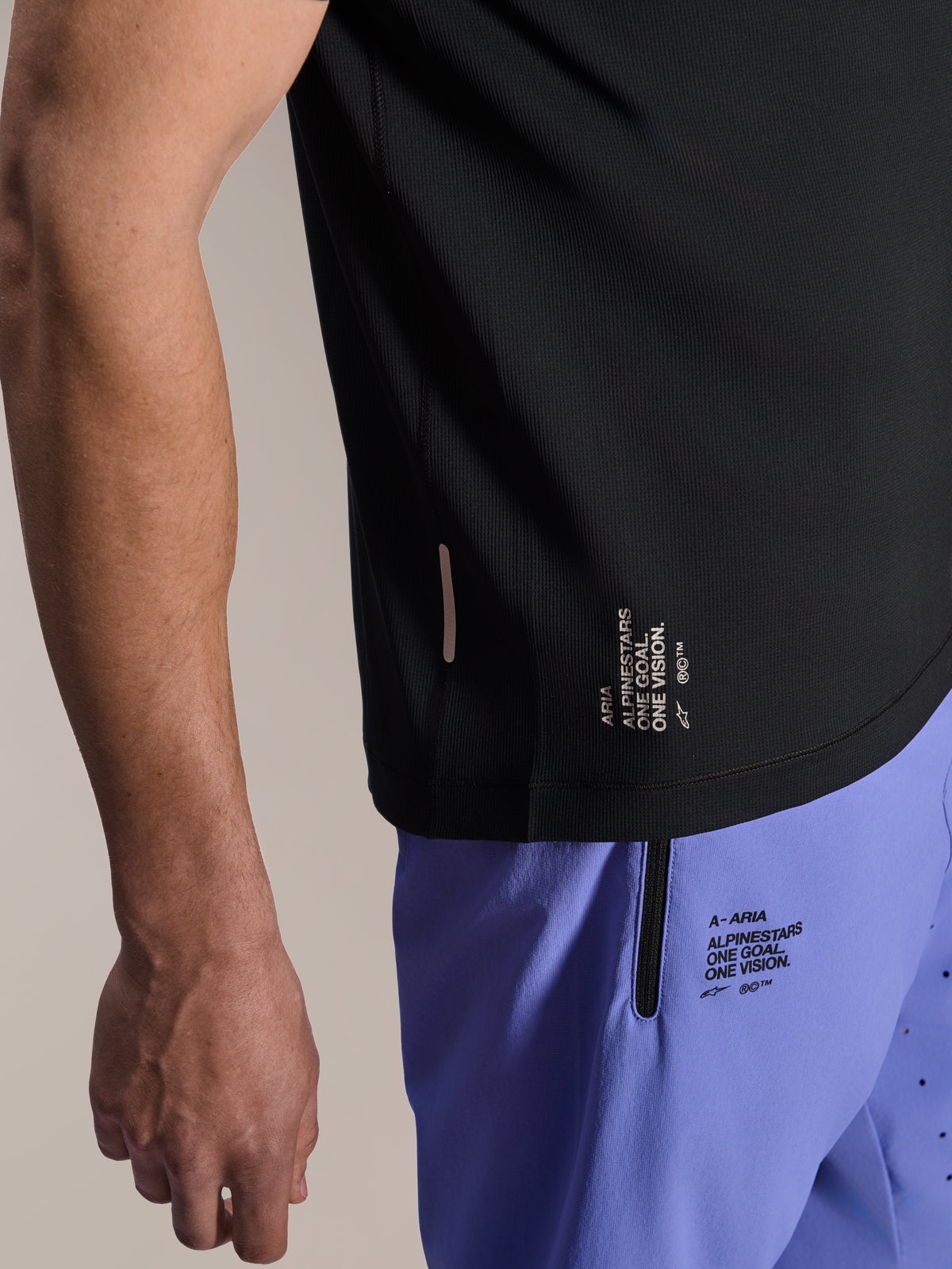 Alpinestars A-Aria Switch Kortærmet Jersey, sort MTB-trøje, nærbillede af sidekant med hvid 'ONE GOAL. ONE VISION.'-branding, båret med lyslilla cykelshorts med matchende logoer
