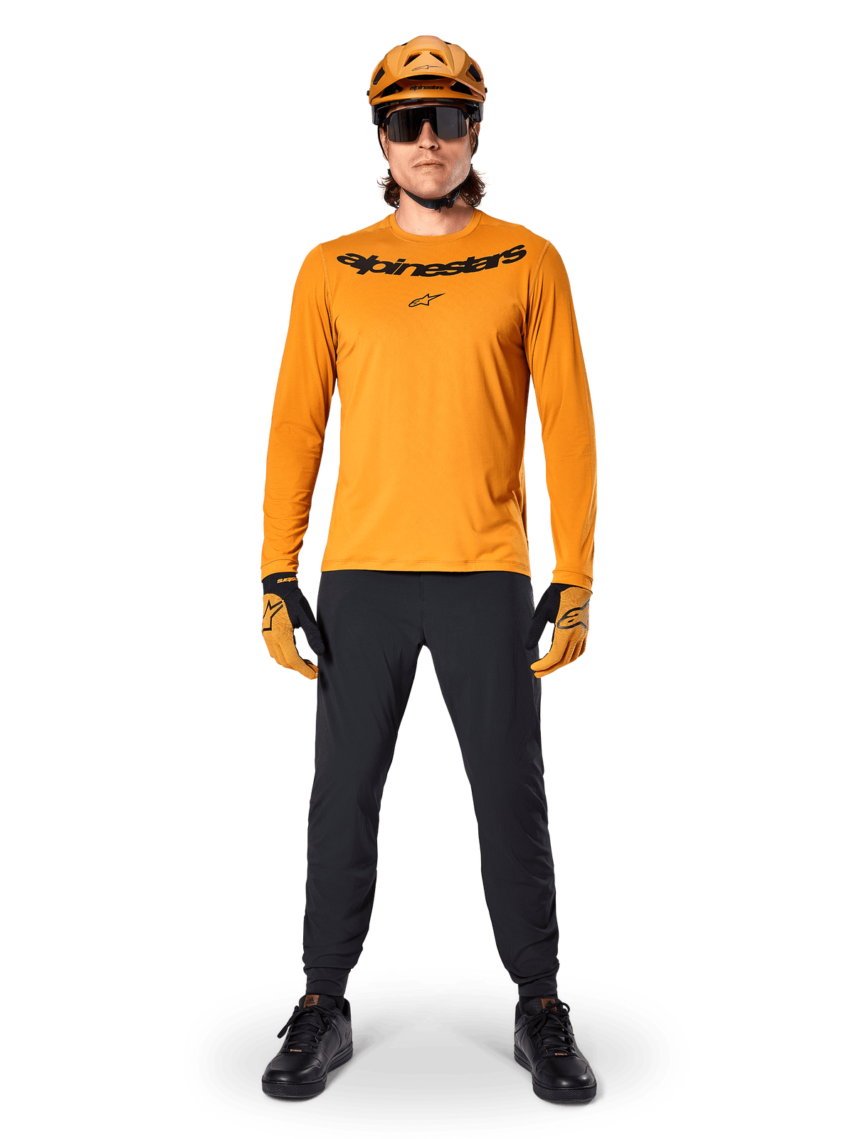 Alpinestars A-Dura Rocker langærmet trøje i Dark Gold, med et sort serigrafiprintet logo på brystet, båret med en matchende guld MTB-hjelm, sorte solbriller og sorte bukser, professionelt mountainbike-udstyr