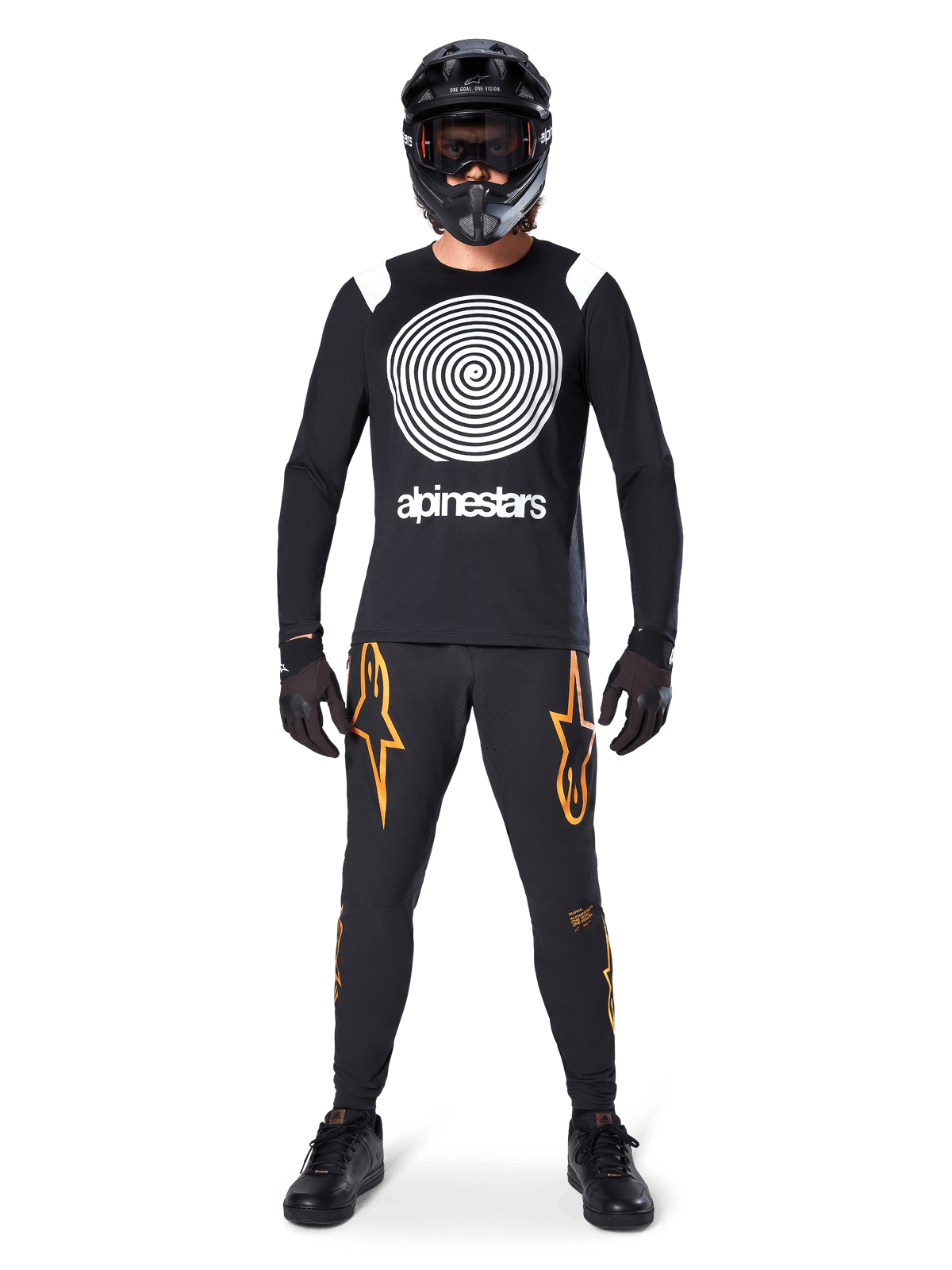 Male model wearing complete mountain bike gear, Alpinestars A-Supra Oscar Long Sleeve Jersey in black with white spiral chest graphic, black pants with orange star logos, black full-face helmet and goggles, black gloves and shoes, coordinated outfit for MTB gravity riding