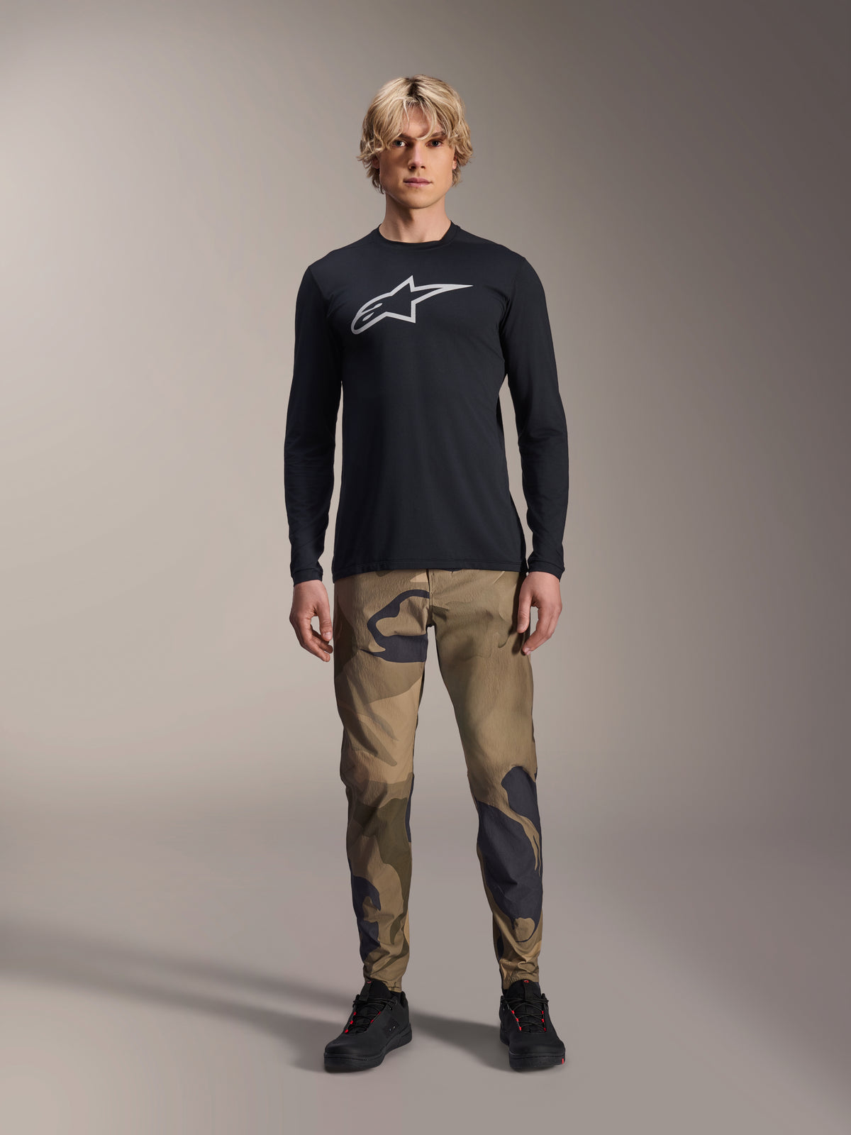 Alpinestars A-Dura Astar langærmet trøje, sort, med et stort hvidt Astars-logo på brystet, båret med camouflagebukser og sorte sko, afslappet mountainbike-performancebeklædning