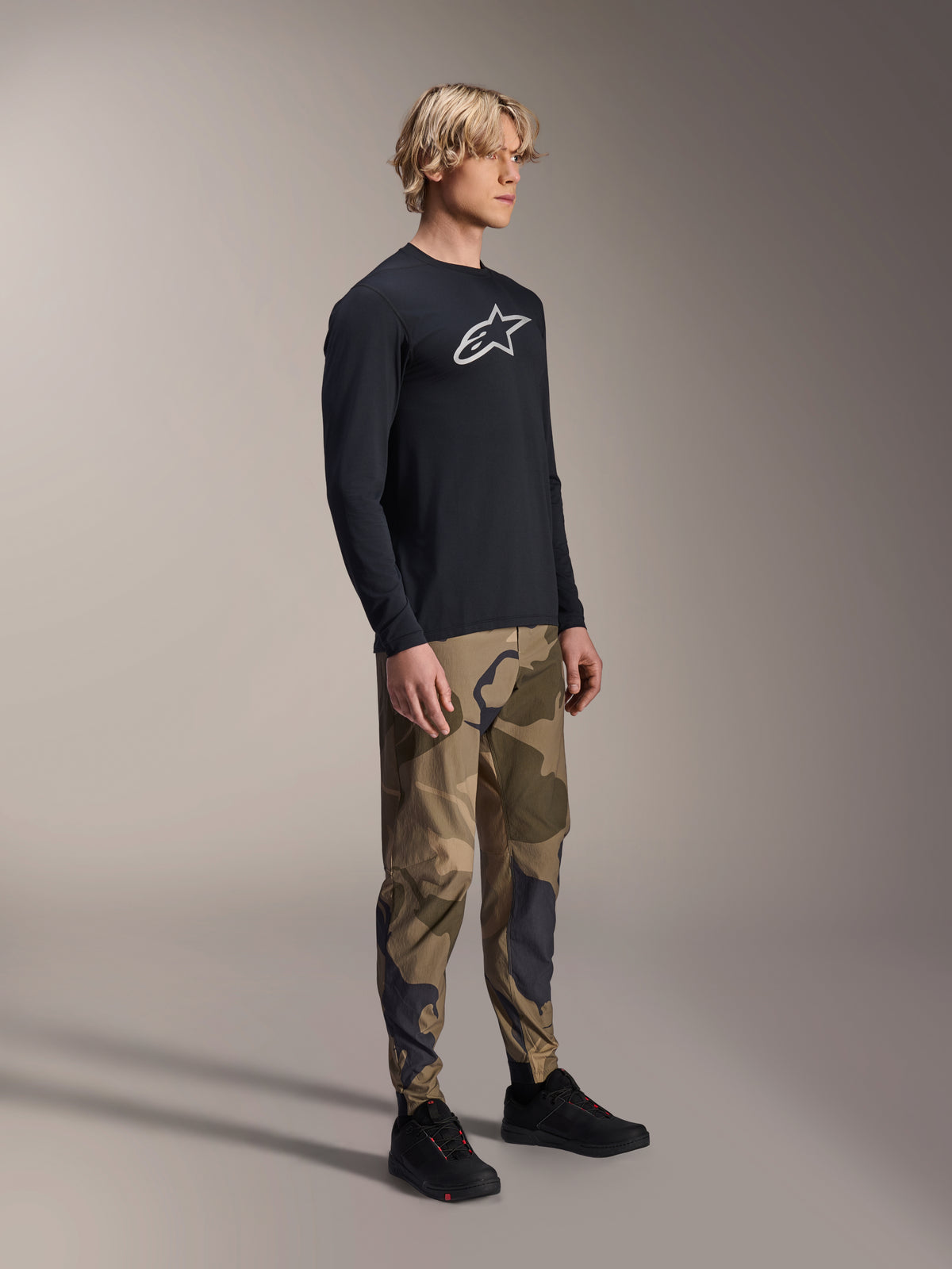 Alpinestars A-Dura Astar langærmet trøje i sort, båret af en model med camouflagebukser og sorte sneakers, med det hvide Astars-logo på brystet, afslappet performance MTB-beklædning til trail-kørsel