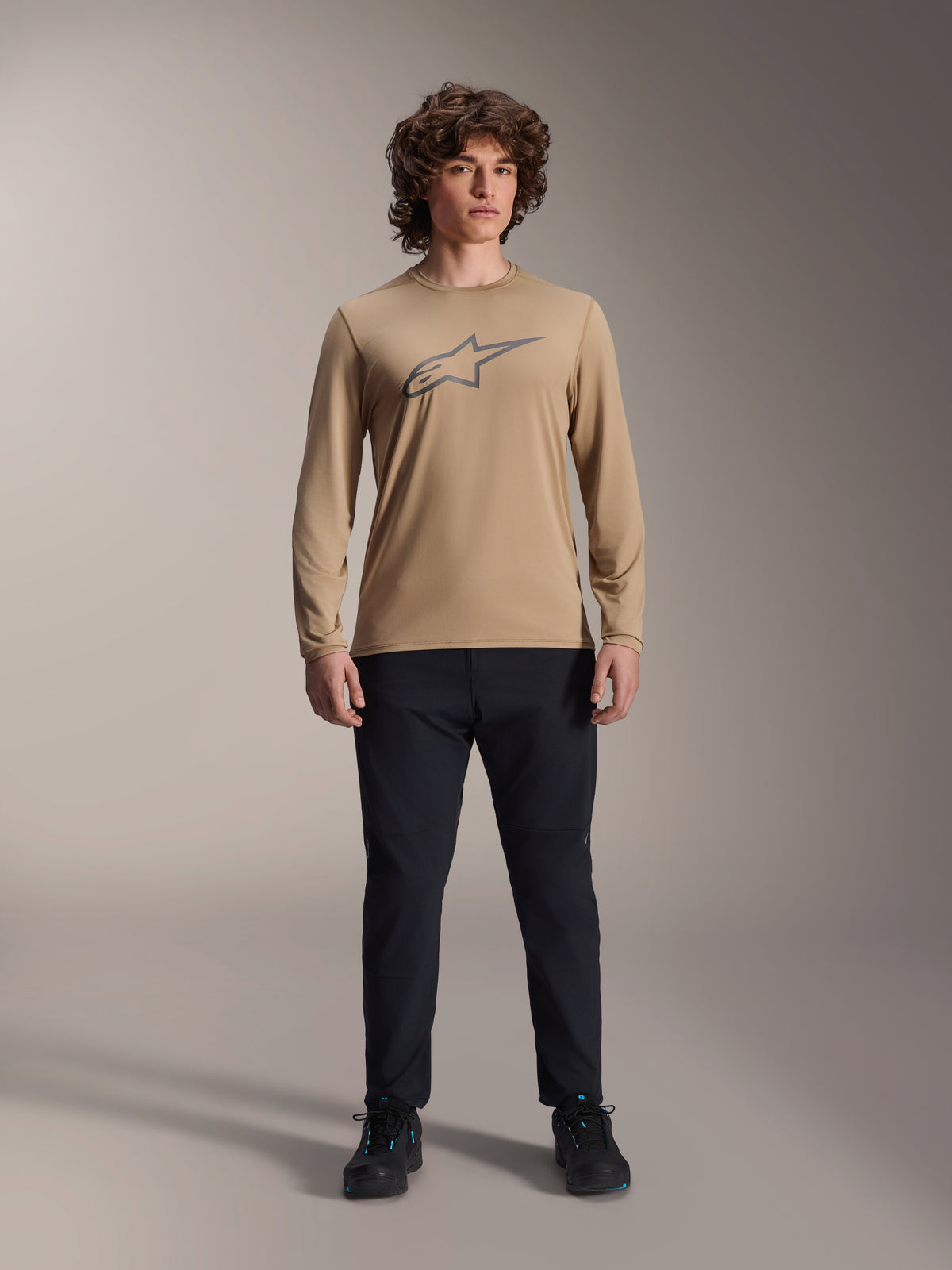 Alpinestars A-Dura Astar langærmet trøje, lysebrun med gråt Astars-logo på brystet, båret af en model med sorte tekniske bukser og sko, afslappet mountainbike-performanceudstyr