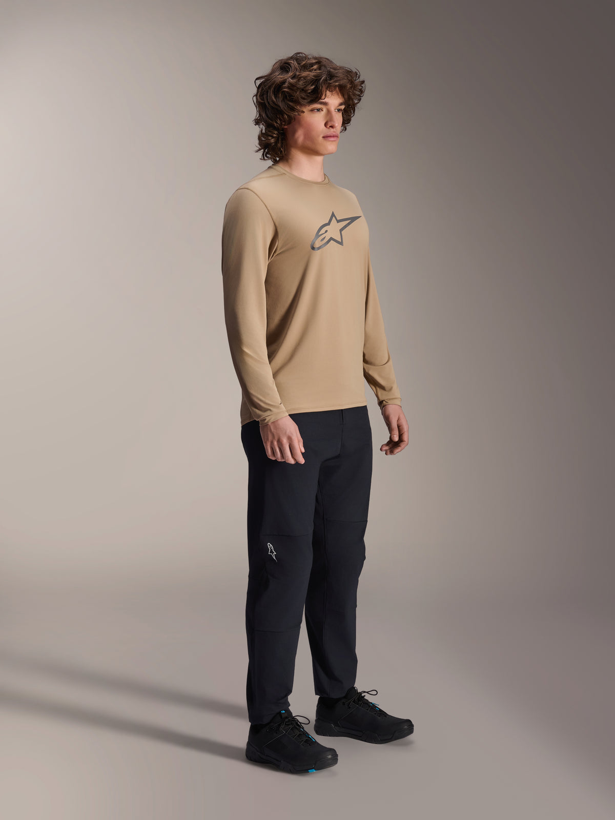 Alpinestars A-Dura Astar langærmet trøje, lysebrun, bæres af en model med sorte bukser og sko, med et gråt Astars-logo på brystet, afslappet mountainbike-performanceudstyr