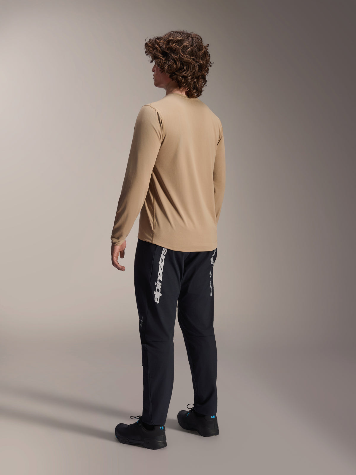 Alpinestars A-Dura Astar langærmet trøje i lys brun, set bagfra på en mandlig model, der viser drop tail-designet, kombineret med sorte bukser og sko, afslappet mountainbike-udstyr.