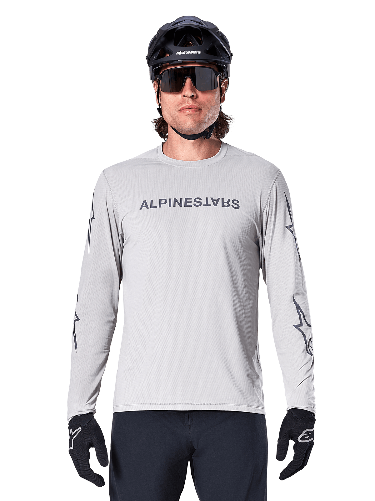 Alpinestars A-Dura Switch langærmet jersey, lysegrå, med mørkegråt tekstlogo på brystet og stjernelogoer på ærmerne, båret af en rytter med sort MTB-hjelm og handsker, funktionelt cykeltøj til trail-kørsel