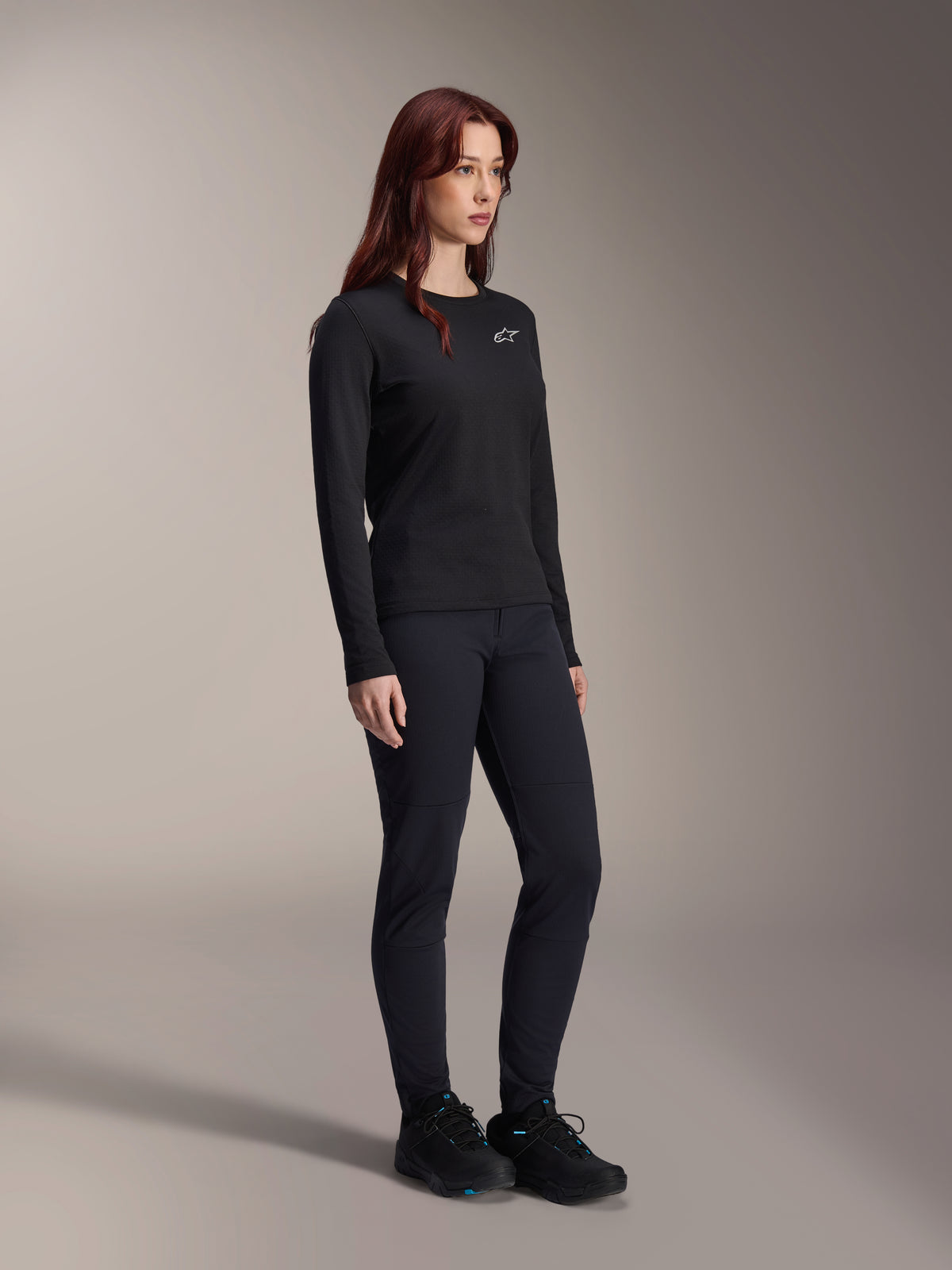Kvinde iført Alpinestars Stella A-Dura Thermal Jersey, langærmet sort MTB-trøje med hvidt logo på brystet, stylet med matchende sorte bukser og sportssko, studioportræt til teknisk cykeltøj