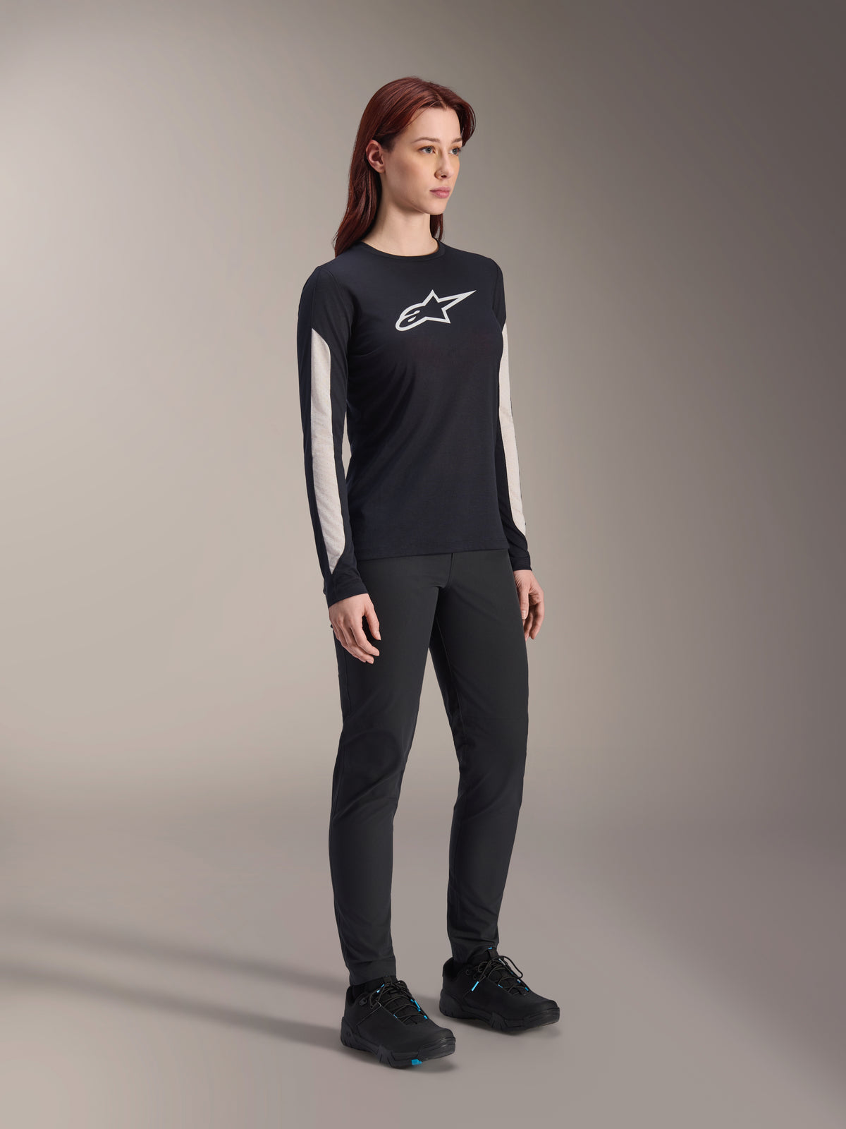 Kvinde iført Alpinestars MTB-udstyr, sort Stella A-Dura Elite Jersey med hvidt brystlogo og hvide ærmepaneler, sorte Stella A-Dura bukser, sorte cykelsko med blå detaljer, komplet performance-outfit til mountainbiking