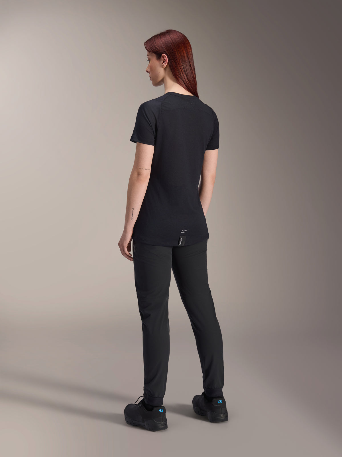 Woman model with red hair wearing the Alpinestars Stella A-Dura Dri Switch Short Sleeve Jersey in Black, rear view, paired with black mountain bike pants and shoes, showing the technical fit and longer drop tail for cycling coverage.