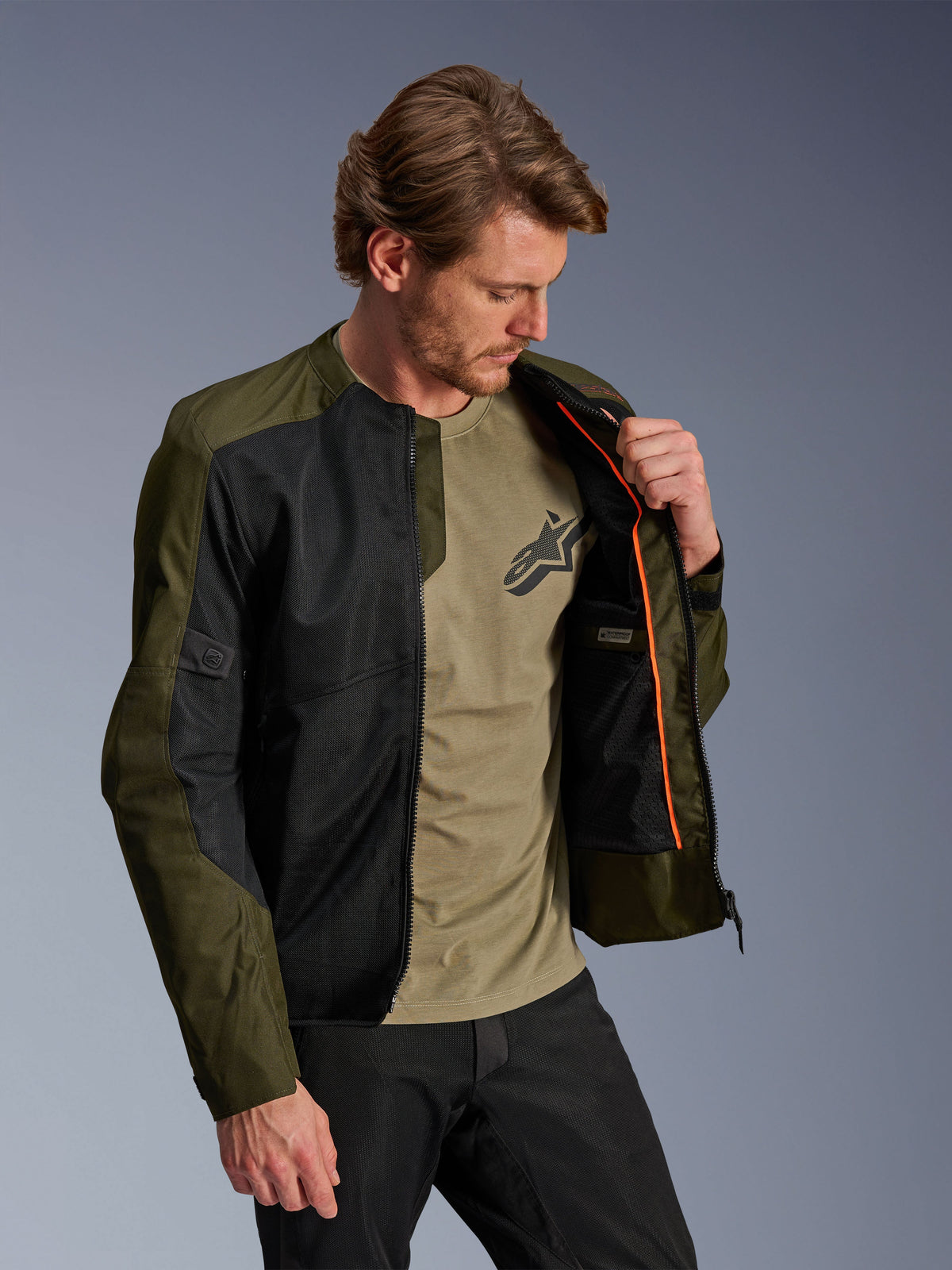 Alpinestars C-1 Air-jakke i khaki og sort, båret af en mandlig model for at vise det orange inderfor og det vandtætte rum, store tech mesh-paneler til luftgennemstrømning, urban moderne motorcykelstil kombineret med en matchende oliven t-shirt og sorte bukser
