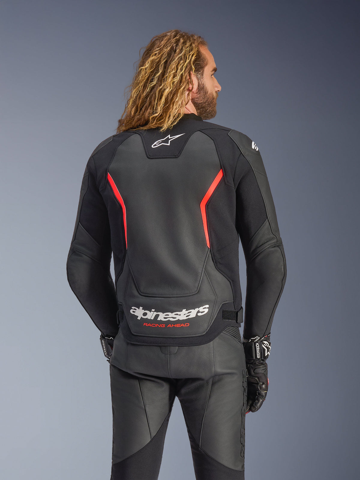 Alpinestars GP Force V2 læderjakke, sort og fluorød, set bagfra på en model, med hvidt Alpinestars-logo og 'RACING AHEAD'-tekst, kombineret med sorte motorcykelbukser og handsker i læder