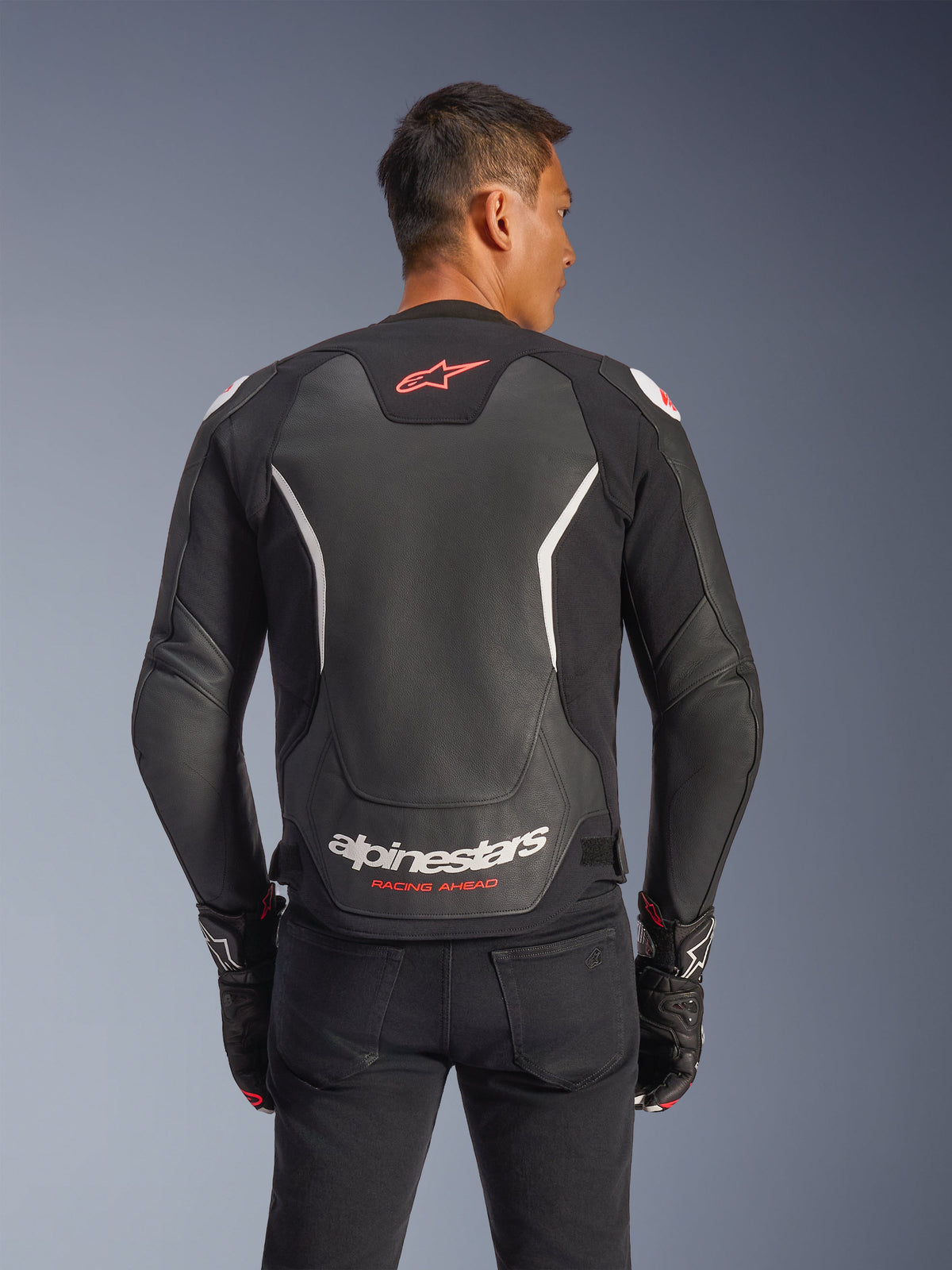 Alpinestars GP Force V2 læderjakke, set bagfra, sort og hvid, med rødt stjernelogo øverst på ryggen og hvid Alpinestars-branding med 'Racing Ahead'-tekst, båret med sorte bukser og matchende handsker, motorcykeljakke i læder med sportspasform