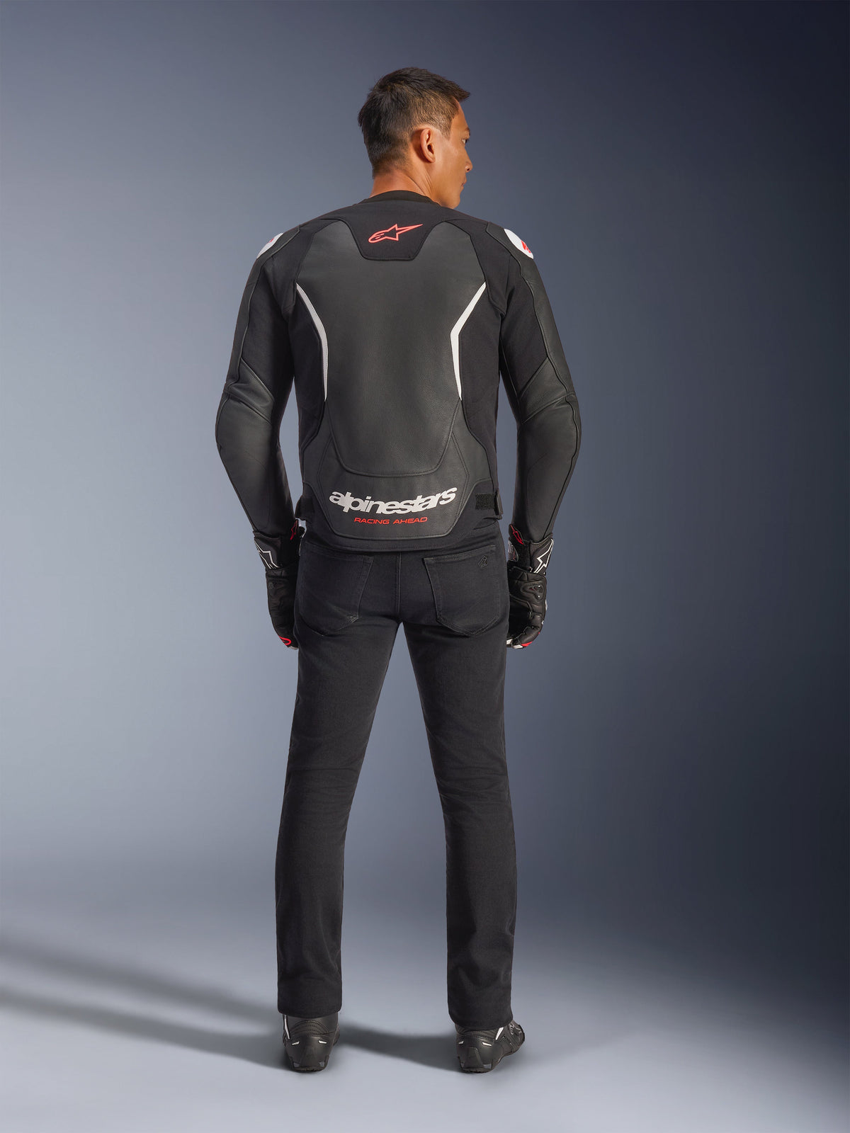 Alpinestars GP Force V2 læderjakke, sort og hvid, set bagfra, båret af en kører med sorte bukser og handsker, med stort hvidt logo på lænden og røde detaljer, premium bovinlæder-konstruktion til motorcykelbrug