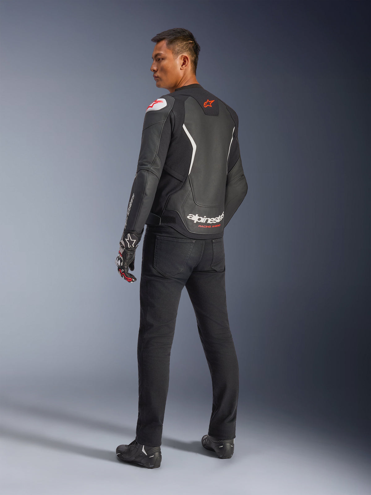 Alpinestars GP Force V2 læderjakke, sort og hvid, set bagfra, viser læderkonstruktion med hvide detaljer og røde logoer, båret med sorte bukser og racinghandsker, med markant Alpinestars-branding på den nederste del af ryggen, motorcykeludstyr med sportspasform