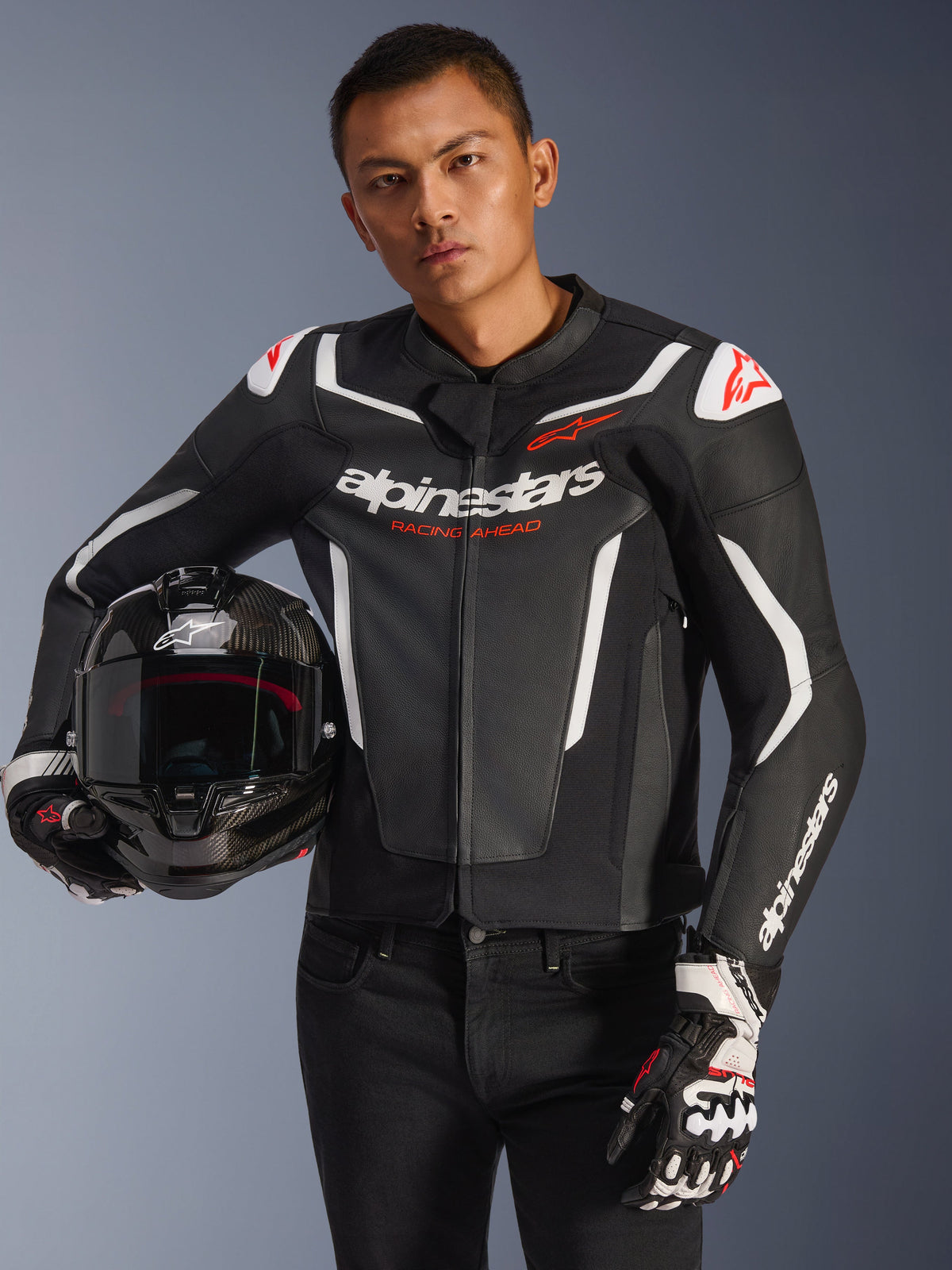 Alpinestars GP Force V2 læderjakke, sort og hvid med røde detaljer, båret af en model der holder en motorcykelhjelm i kulfiber og har racerhandsker på, med hvide kontrastpaneler og Alpinestars-branding.