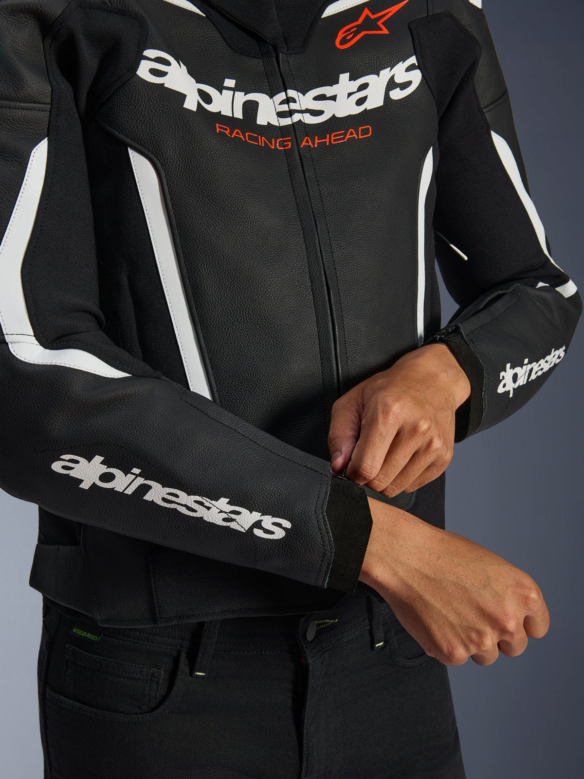 Alpinestars GP Force V2 læderjakke, sort og hvid, nærbillede af kører der justerer lynlåsen ved manchetten, hvid branding på bryst og ærmer med orange 'Racing Ahead' tekst, premium bovinlæder til sportslig motorcykelkørsel