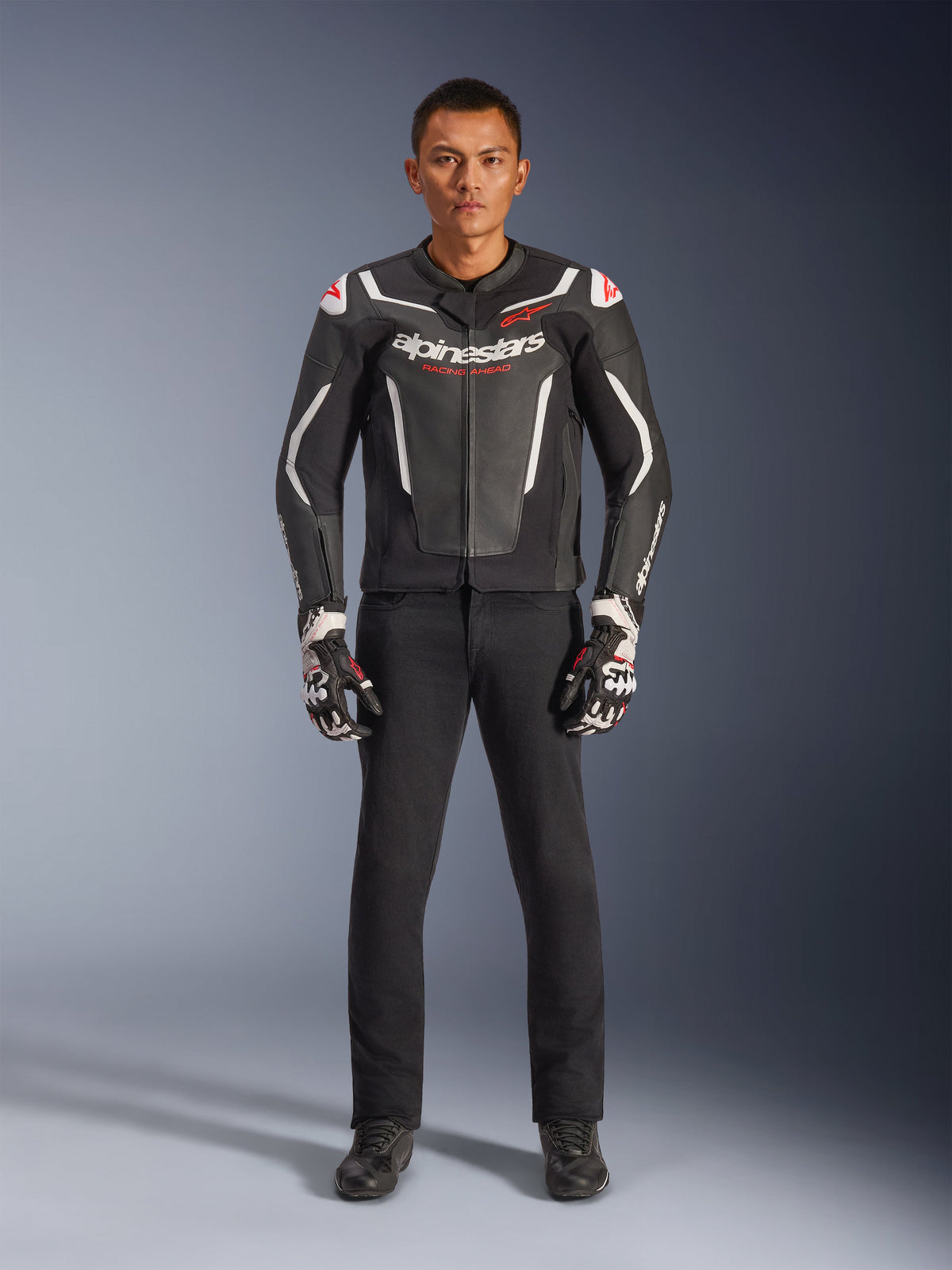 Mandlig model iført Alpinestars GP Force V2 læderjakke i sort og hvid, kombineret med sorte motorcykelbukser og sort-hvide handsker, med fremtrædende Alpinestars-branding og røde skulderdetaljer, professionelt sportsmotorcykeludstyr
