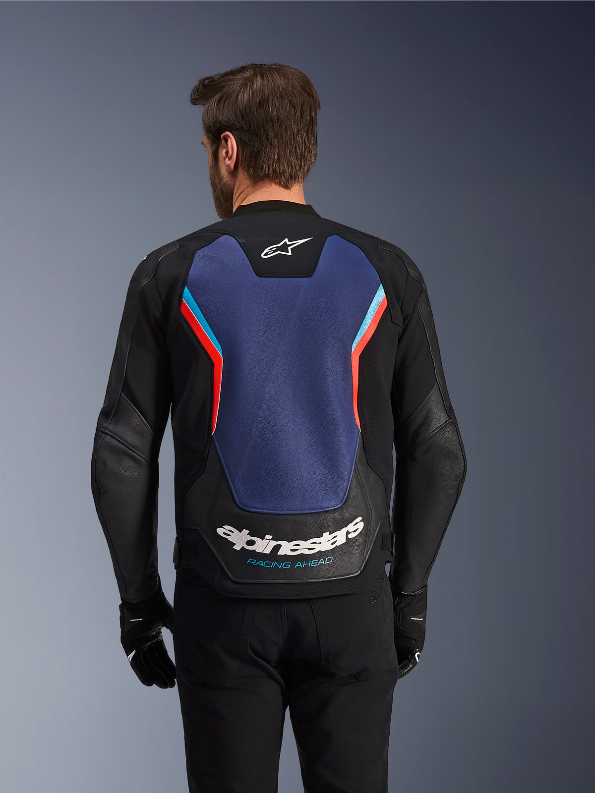 Set bagfra af Alpinestars GP Force V2 læderjakken i Black, Blue og Red Fluo, med et stort blåt rygpanel med fluorescerende røde og lyseblå detaljer, hvidt Alpinestars-logo og 'Racing Ahead'-branding på den nederste del af ryggen, båret af en kører med sorte handsker og bukser.