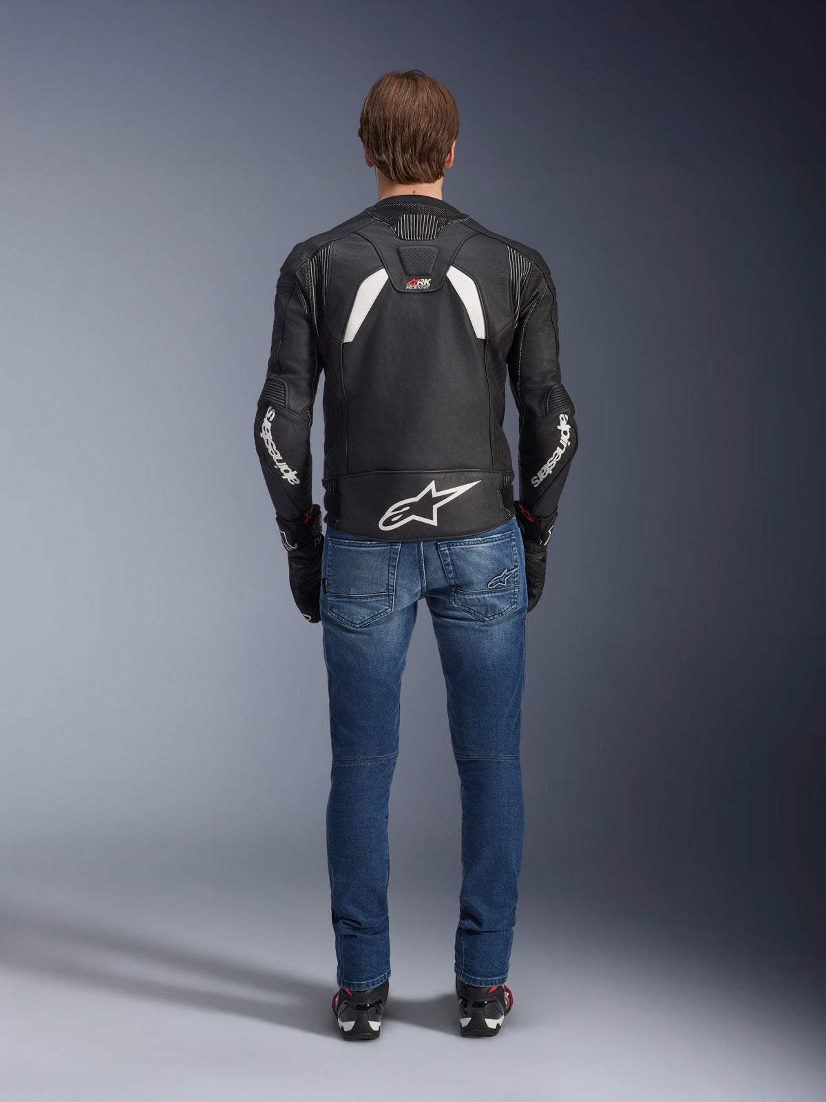 Rear view of the Alpinestars GP Plus R V4 Rideknit Leather Jacket, Black and White, featuring a large white logo on the lower back and branding on the sleeves, worn with blue riding jeans and black boots, highlighting the technical construction and sport fit.