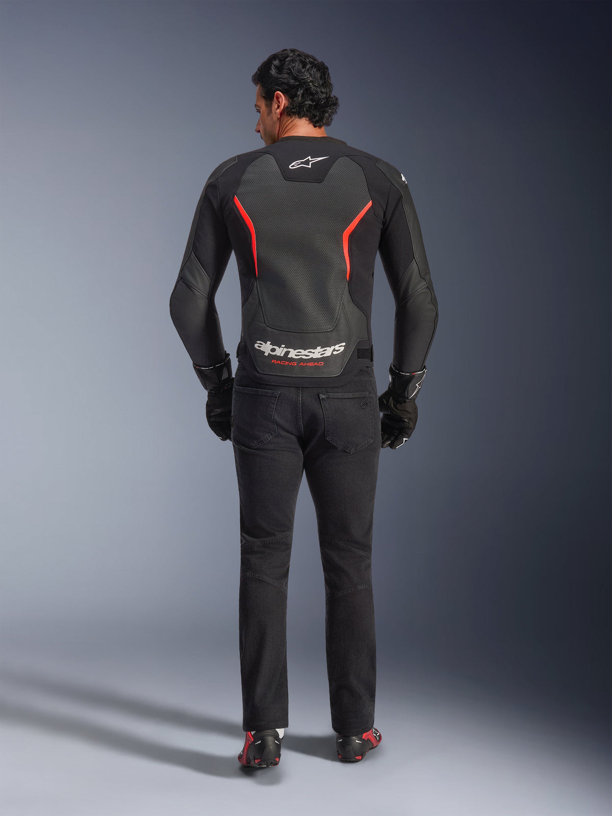 Alpinestars GP Force V2 Air læderjakke i Black Red Fluo, set bagfra på model, med perforerede læderpaneler på ryggen, røde detaljer og Alpinestars-branding, kombineret med sorte kørejeans og handsker for et sporty motorcykel-look.
