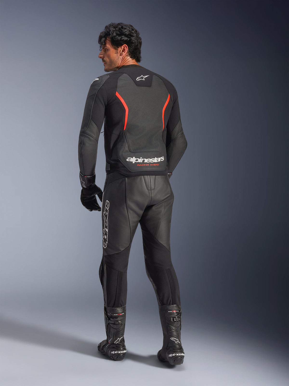 Bagfra-visning af en model iført Alpinestars GP Force V2 Air læderjakke i Black Red Fluo, med perforerede læderpaneler og markant Alpinestars-branding på lænden, stylet med matchende sorte motorcykelbukser og støvler i læder.