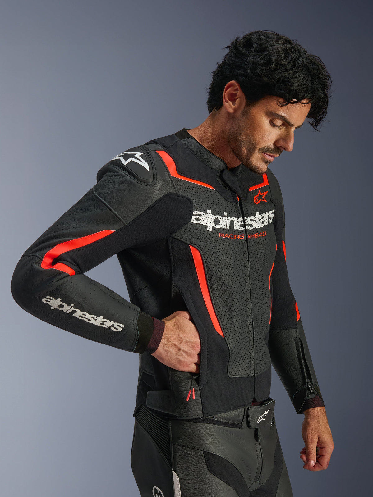 Alpinestars GP Force V2 Air læderjakke, sort og rød fluorescerende, perforeret læder motorcykeljakke, model iført jakken rækker ind i en sidelomme med lynlås, med fluorescerende røde detaljer og hvidt Alpinestars-logo, professionelt sportskøreudstyr.