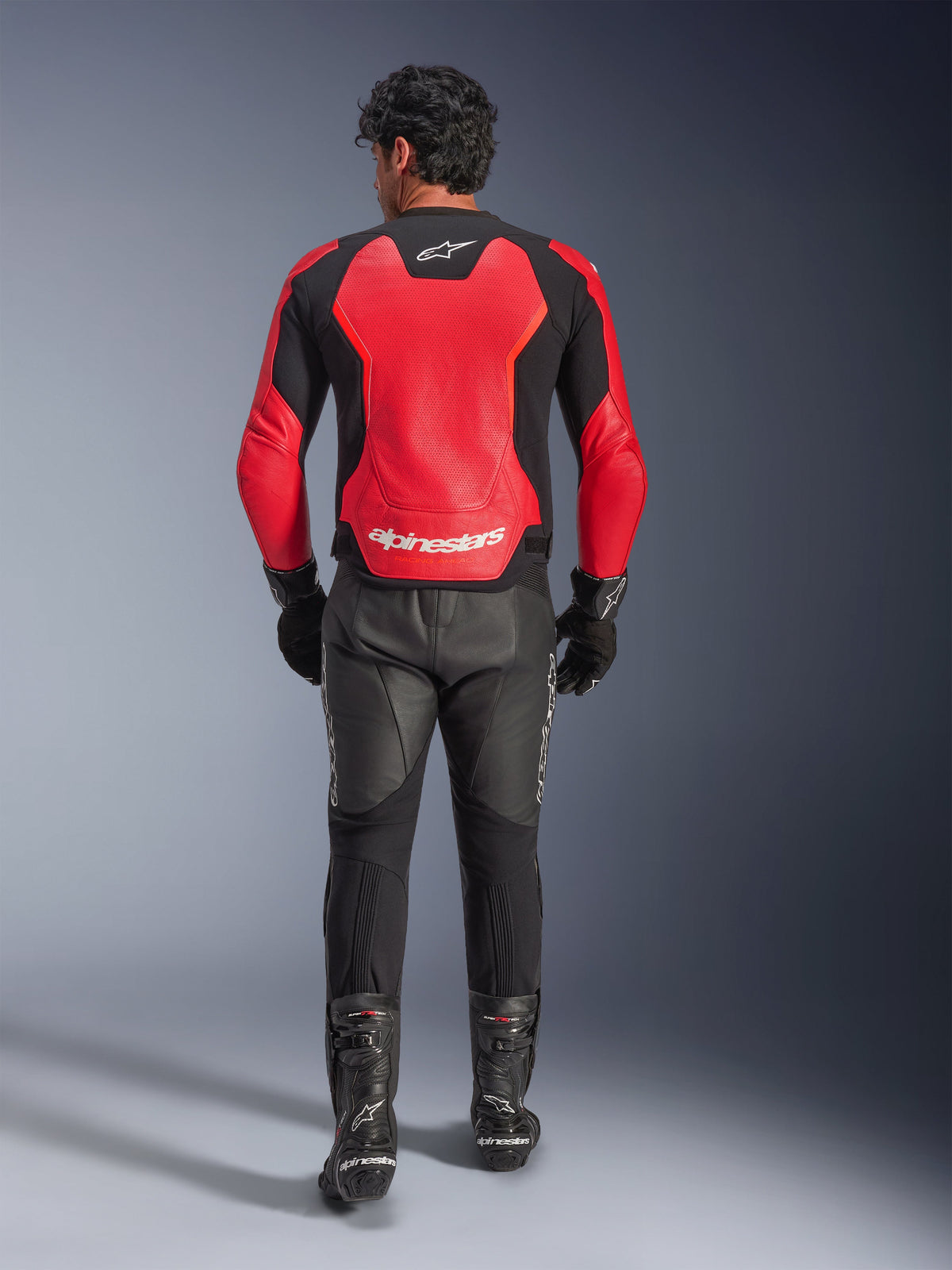 Rear view of a rider wearing the Alpinestars GP Force V2 Air Leather Jacket in Red and Black, featuring large perforated panels and white Alpinestars logo on the back, paired with black leather motorcycle pants and racing boots.
