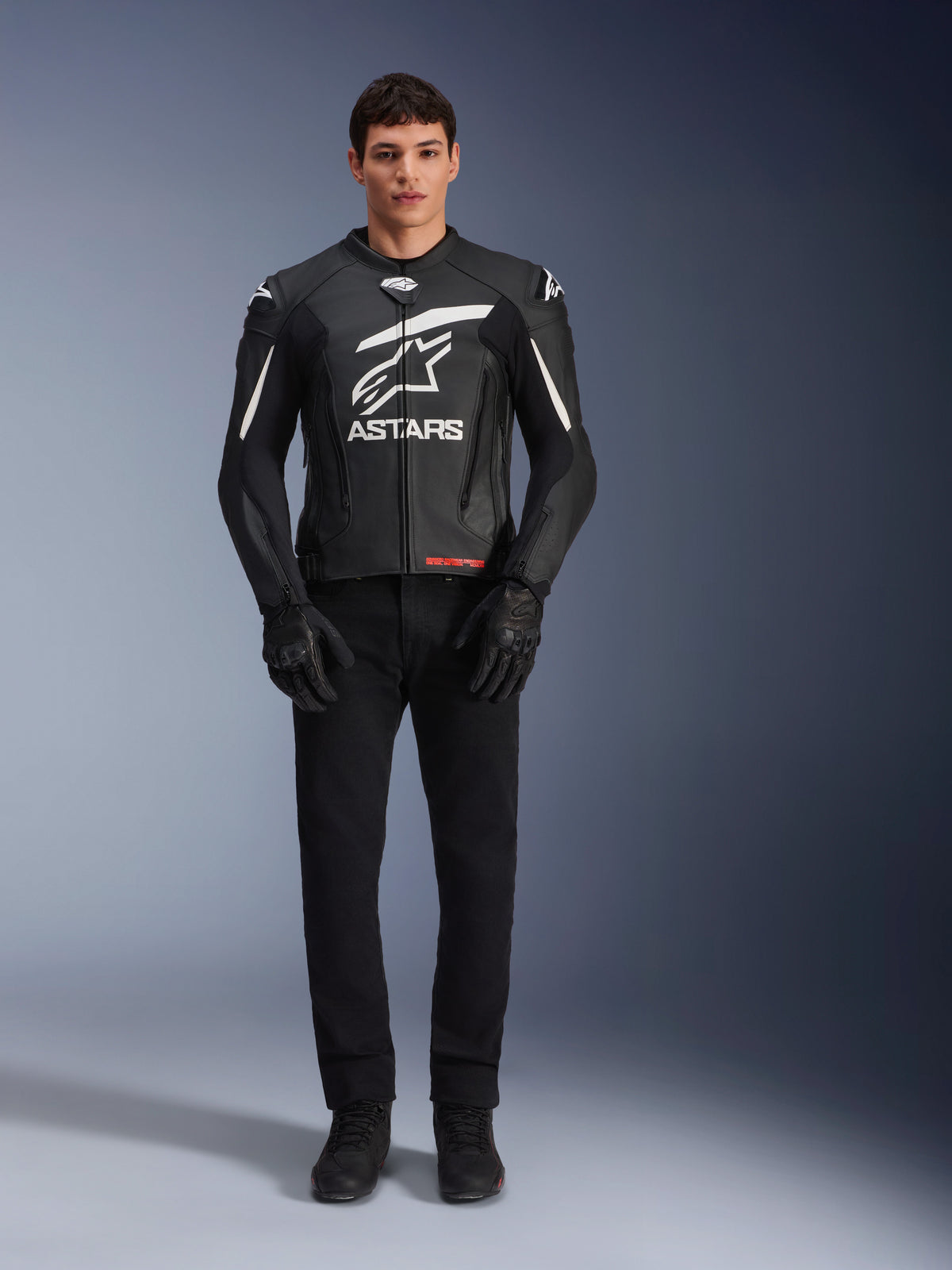 Male model wearing Alpinestars GP Plus V4 Leather Jacket, Black and White, featuring bold white Astars branding on the chest and white accents on the shoulders, paired with black riding gloves and pants, sleek sport-oriented motorcycle apparel