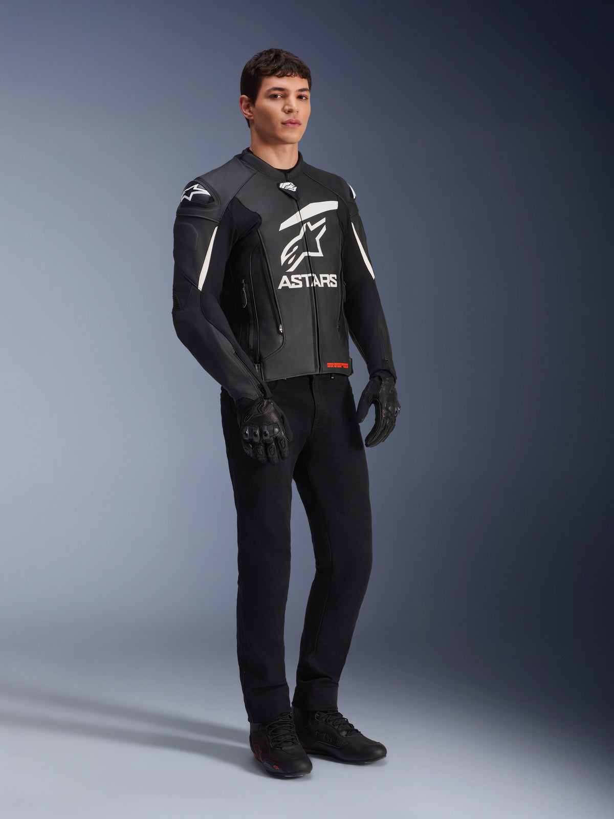 Alpinestars GP Plus V4 Leather Jacket, Sport Motorcycle Jacket, Black and White, worn by a male model with black riding pants and gloves, featuring a large white ASTARS logo on the chest and external DFS shoulder sliders, professional racing-derived design