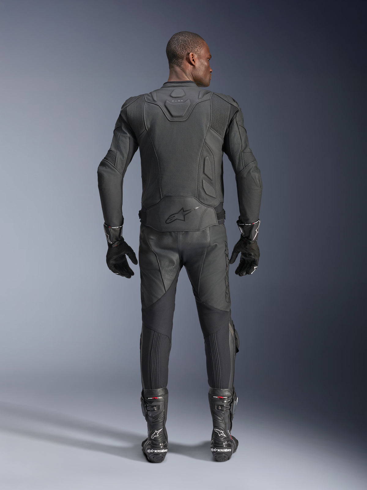 Mandlig model set bagfra iført Alpinestars Dusk Airflow læderjakke i sort, kombineret med matchende sorte læderracerbukser, handsker og støvler, med omfattende perforeringer for ventilation og Alpinestars-logo på lænden.