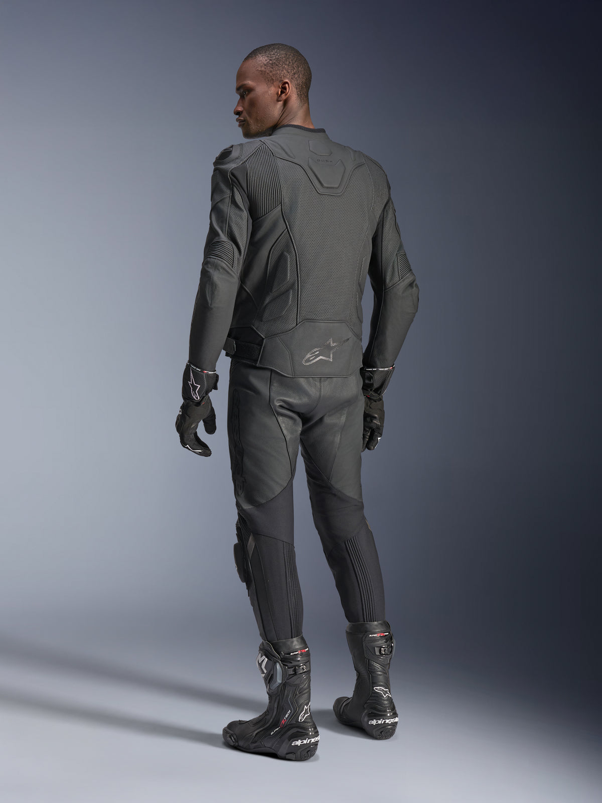 Mandlig motorcyklist set bagfra iført Alpinestars Dusk Airflow læderjakken i Black Black, kombineret med matchende læderbukser, handsker og støvler, hvilket fremhæver de omfattende perforeringer på ryggen og den sportslige pasform.