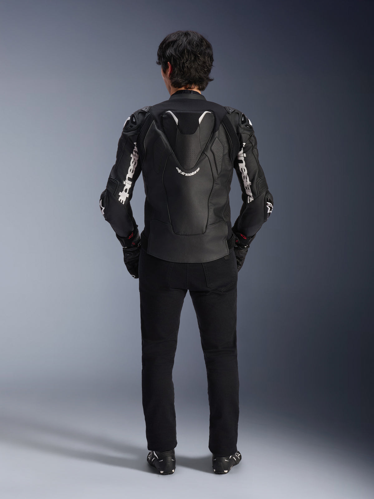 Alpinestars Atem V5 Leather Jacket, Motorcycle Racing Jacket, Black and White, rear view on rider, featuring aerodynamic speed hump and white Alpinestars branding on sleeves and back, technical leather construction, worn with black pants and riding boots