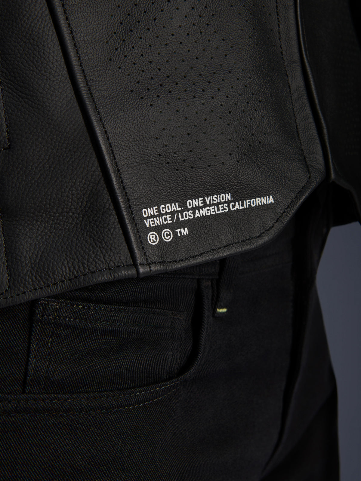 Close-up of the Alpinestars Atem V5 Leather Jacket in Black, featuring the lower back panel with 'ONE GOAL. ONE VISION. VENICE / LOS ANGELES CALIFORNIA' branding and perforated leather texture.
