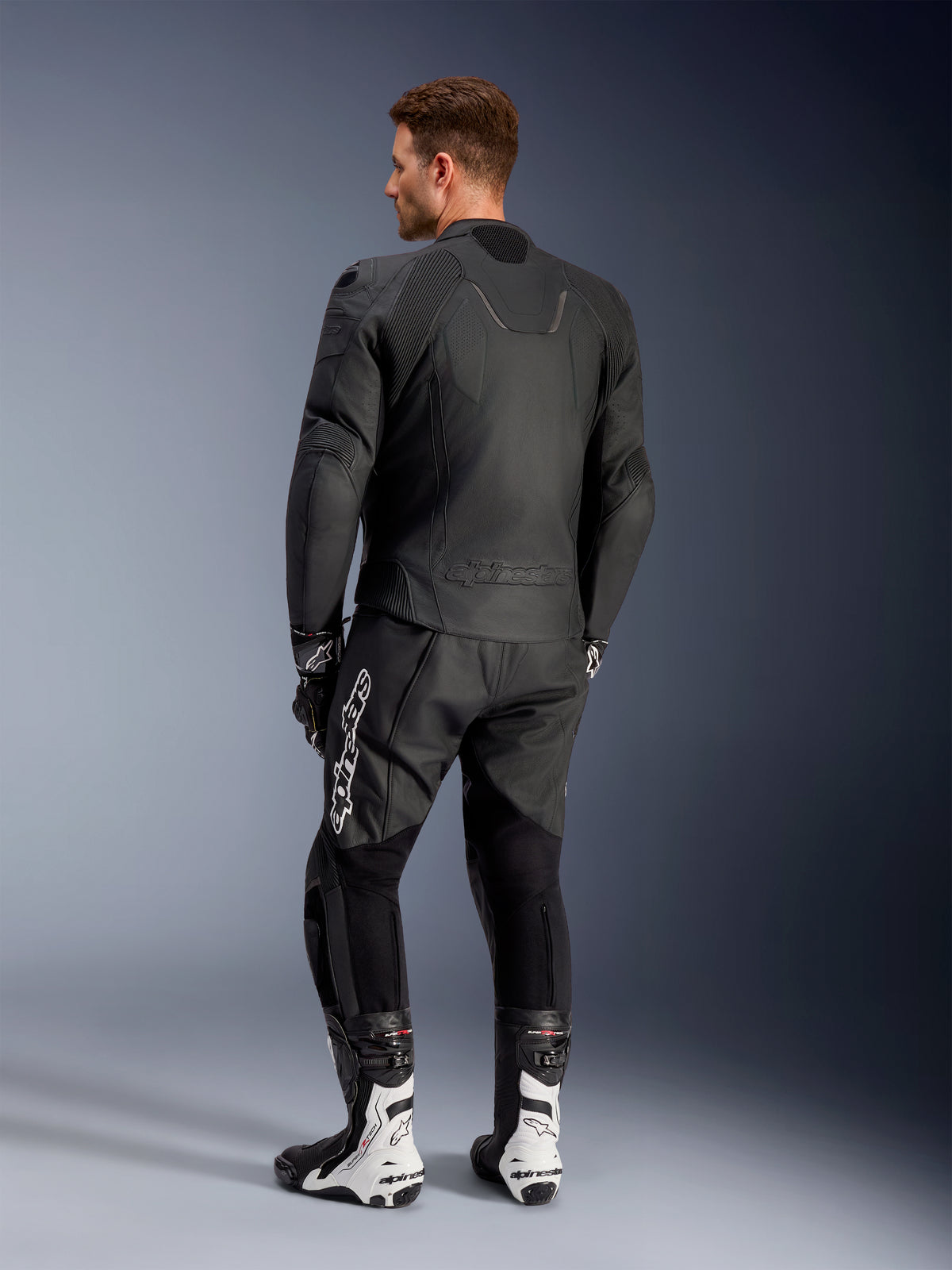 Alpinestars Caliber læderjakke, sort motorcykeljakke til vejbrug, set bagfra, med perforeret læder og akkordeon-strækindsatser ved taljen og skuldrene, præget logo, båret med sorte læderbukser og hvide racingstøvler