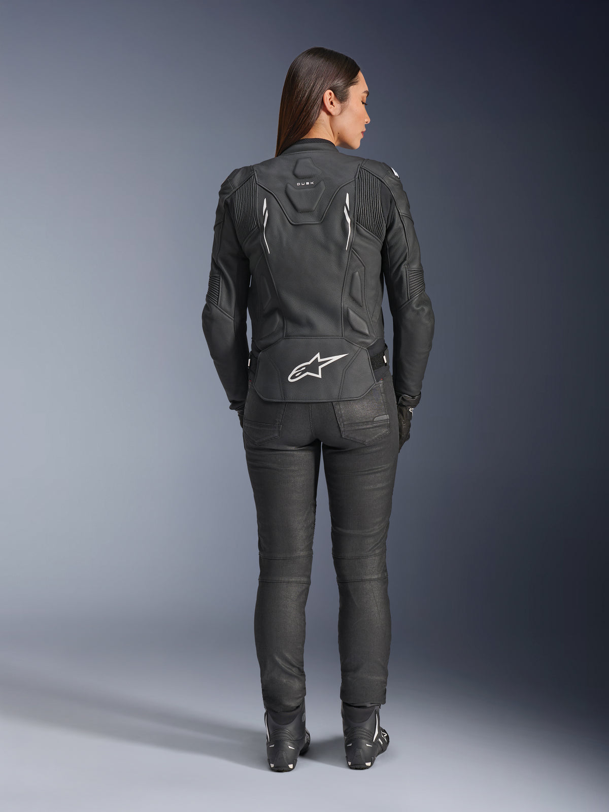 Alpinestars Stella Dusk læderjakke til kvinder, sort, set bagfra med hvidt Alpinestars-logo på den nederste del af ryggen og 'DUSK'-branding øverst, båret med sorte kørebukser og støvler, hvilket viser den konturerede kvindespecifikke pasform og beskyttende læderkonstruktion.