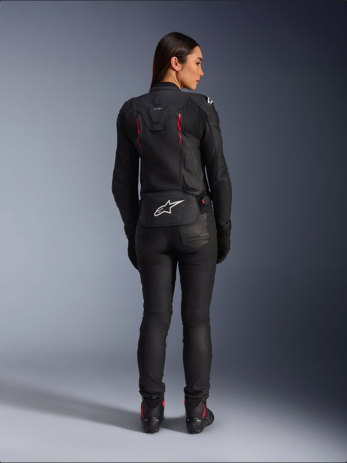 Alpinestars Stella Dusk læderjakke, set bagfra, sort læder med Diva Pink og hvide detaljer, hvidt Alpinestars-logo nederst på ryggen og lyserøde ventilationsdetaljer, båret med sorte kørebukser og støvler, premium sportsmotorcykeludstyr til kvinder