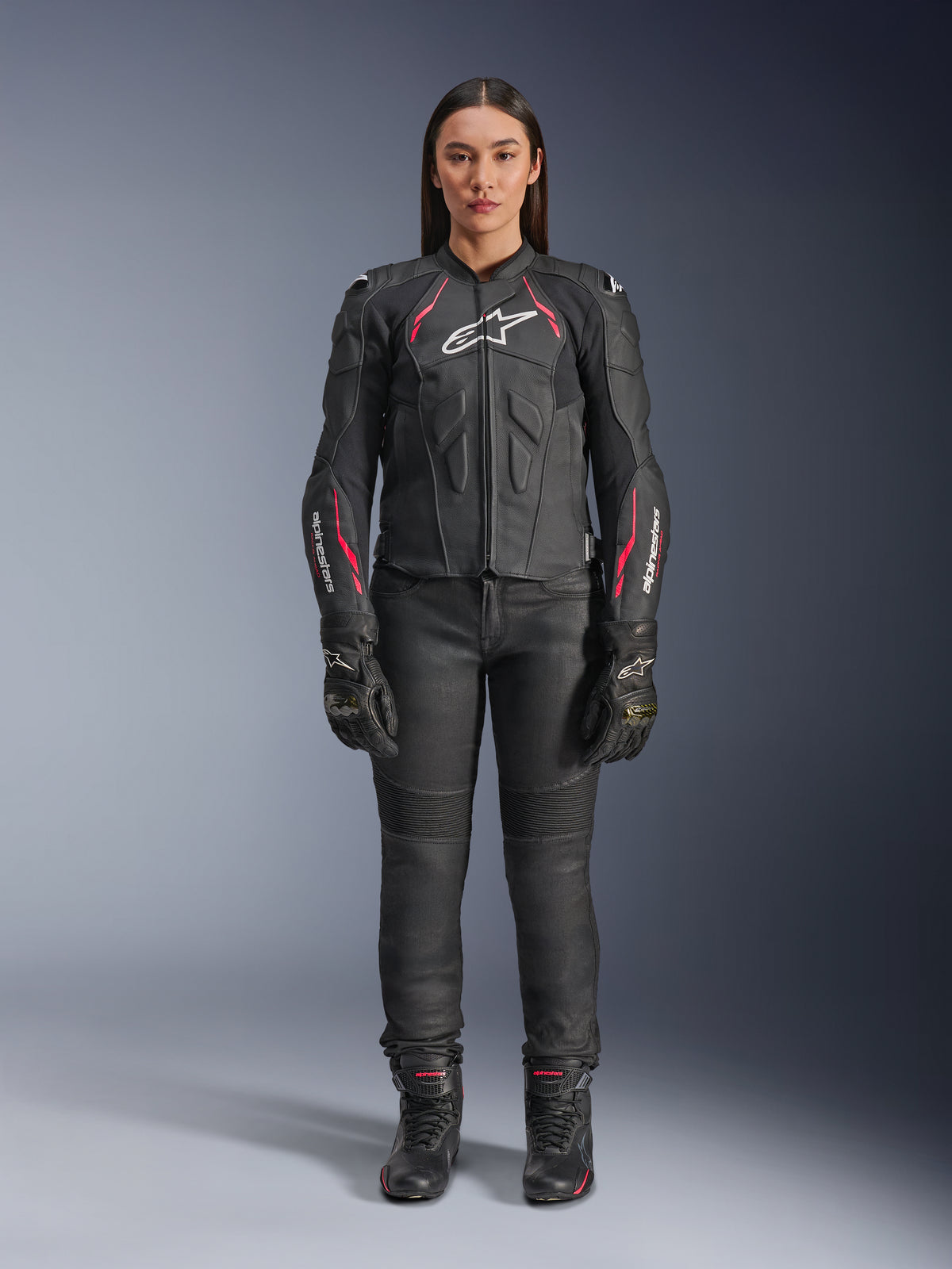 En kvinde iført Alpinestars Stella Dusk læderjakke i Black Diva Pink White, med hvidt logo på brystet og lyserøde detaljer på skuldre og ærmer, kombineret med sorte motorcykelbukser, handsker og støvler.