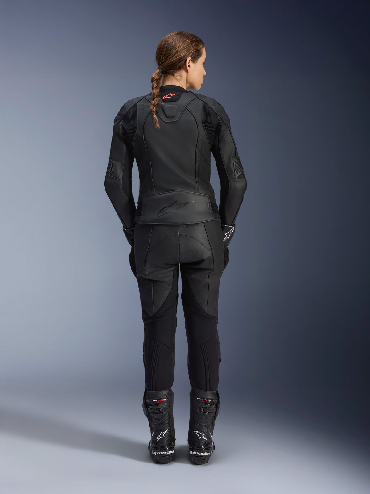 Alpinestars Stella GP Plus V4 læderjakke til kvinder, Black Black, fuldkropsvisning bagfra af en kvindelig rytter iført matchende læderbukser og støvler, strømlinet minimalistisk design med et lille rødt logo i nakken, skræddersyet sports-silhuet til kvinder