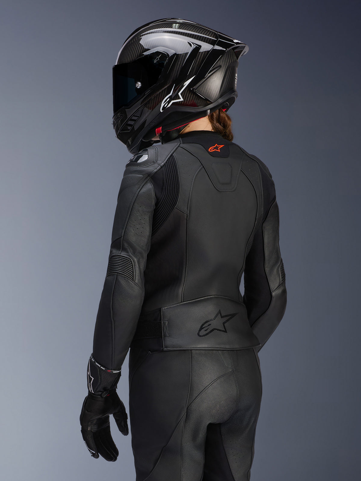 Kvindelig rytter set bagfra iført Alpinestars Stella GP Plus V4 læderjakke til kvinder i sort, matchende læderbukser og handsker samt en sort full-face hjelm i carbon-stil, der fremhæver jakkens aerodynamiske pasform og prægede logo på den nederste del af ryggen.
