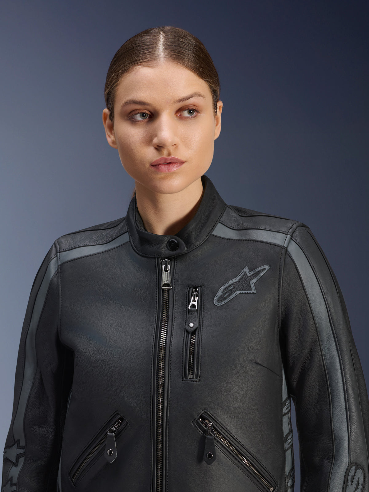 Kvindelig model iført Alpinestars Stella Dyno læderjakke i Black Tar Gray Dark Gray, premium motorcykeljakke i bovinelæder med et heritage-design, grå skulderdetaljer og flere lynlåslommer, skræddersyet til en kvindespecifik pasform.