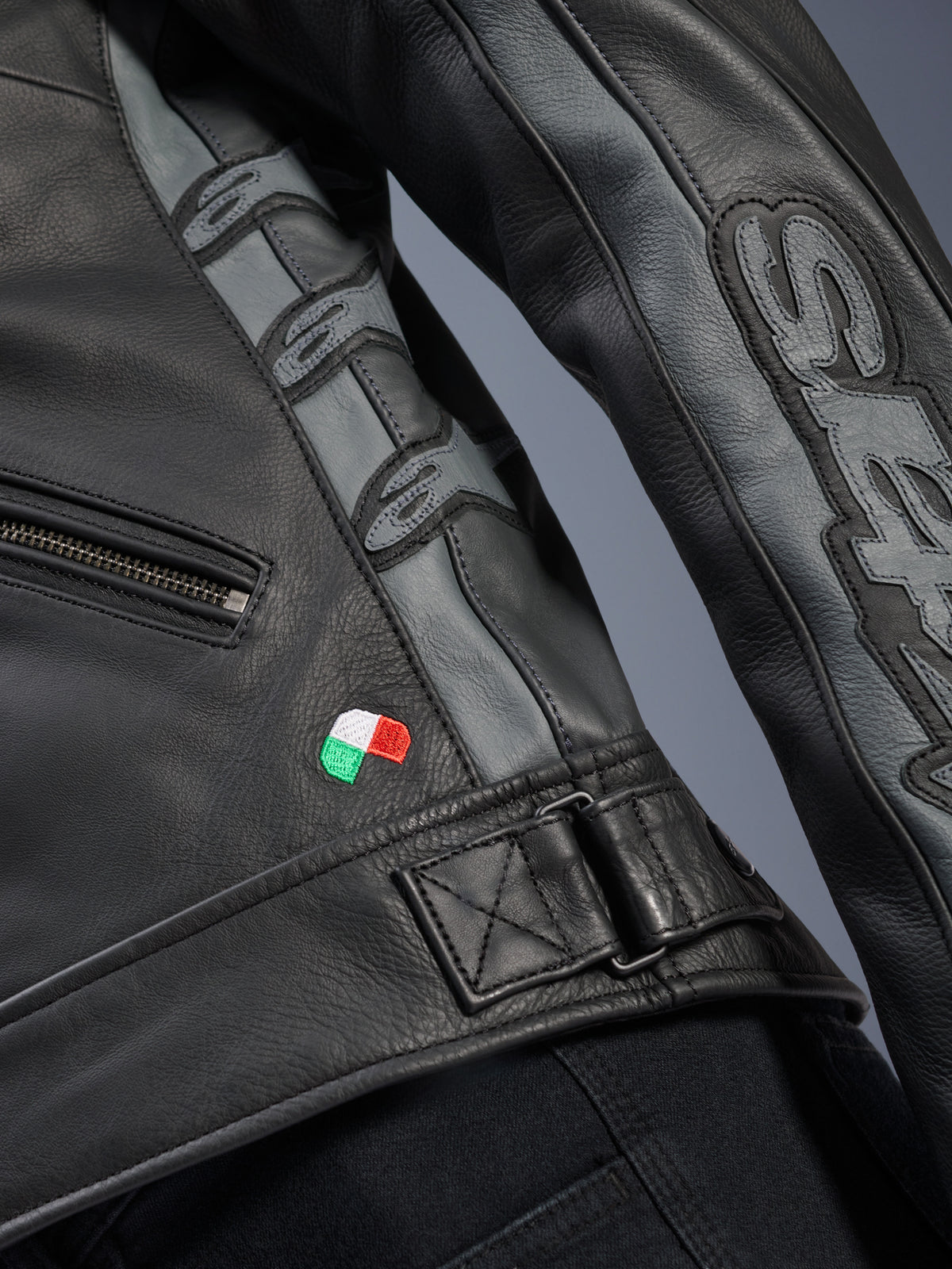 Alpinestars Stella Dyno læderjakke detalje, sort og Tar Gray bovinelæder, nærbillede med lille broderet italiensk flag, justerbart taljespænde og grå læderpaneler med prægede Alpinestars-logoer og Stella-branding på ærmet.