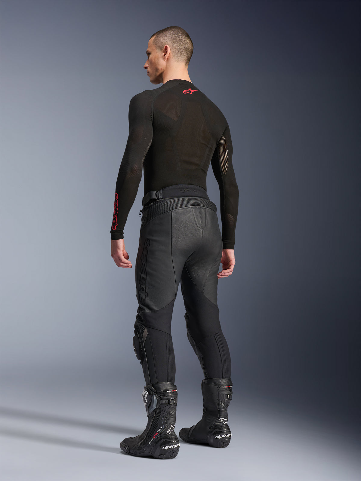 Alpinestars Missile V3 læderbukser, sort, set bagfra på model, båret med et sort langærmet basislag og sorte motorcykelstøvler, med akkordeon-læderstrækpaneler og et forbøjet design for en ergonomisk kørepasform.