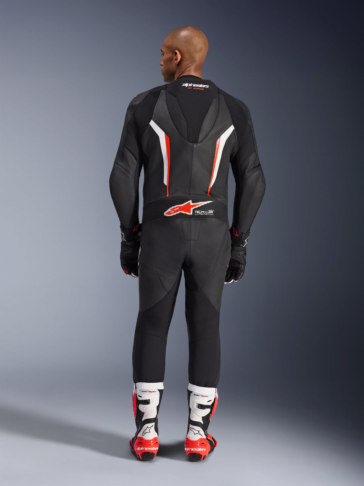 Alpinestars GP Force V2 1-delt læderdragt set bagfra, sort, hvid og rød fluo, fremhæver den aerodynamiske pukkel og Tech-Air Ready-branding, kombineret med hvide og røde professionelle racingstøvler og sorte handsker.
