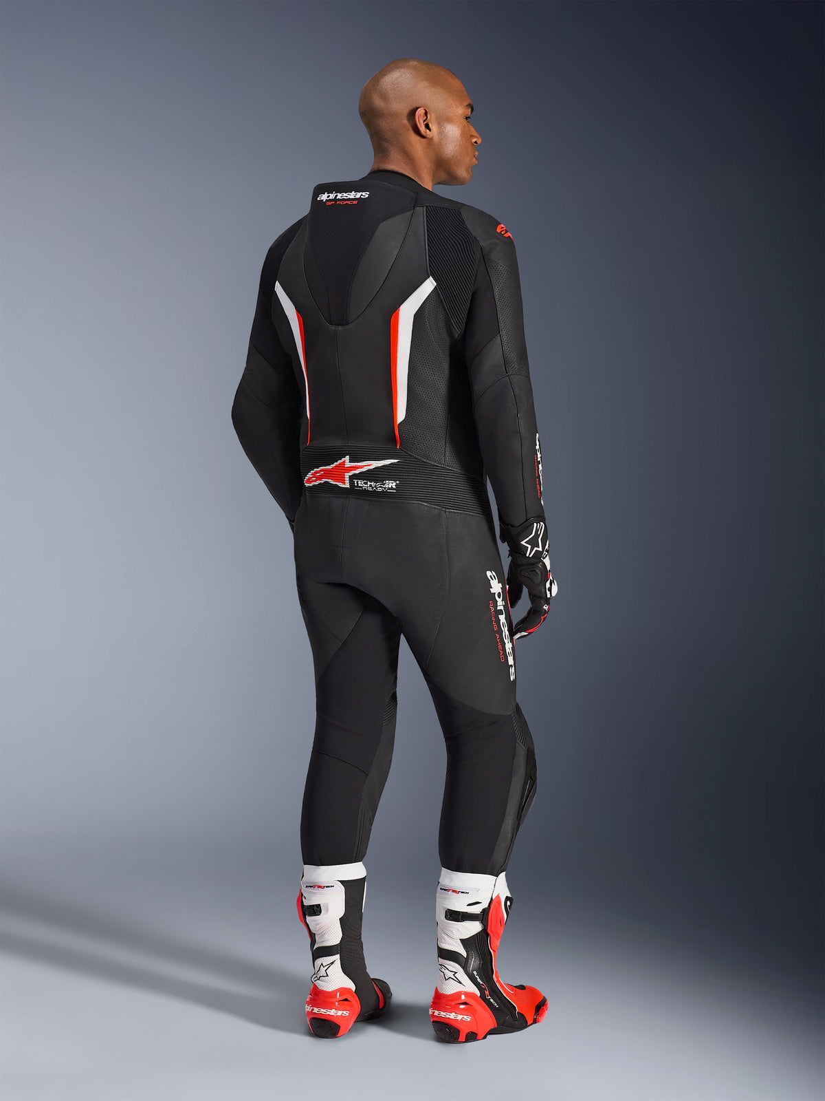 Kører iført GP Force V2 læderdragt i ét stykke, sort med hvide og røde fluo-detaljer, set bagfra, som viser den aerodynamiske pukkel og Alpinestars-branding, kombineret med matchende racerhandsker og hvide og røde performance-støvler, professionelt motorcykelracerudstyr