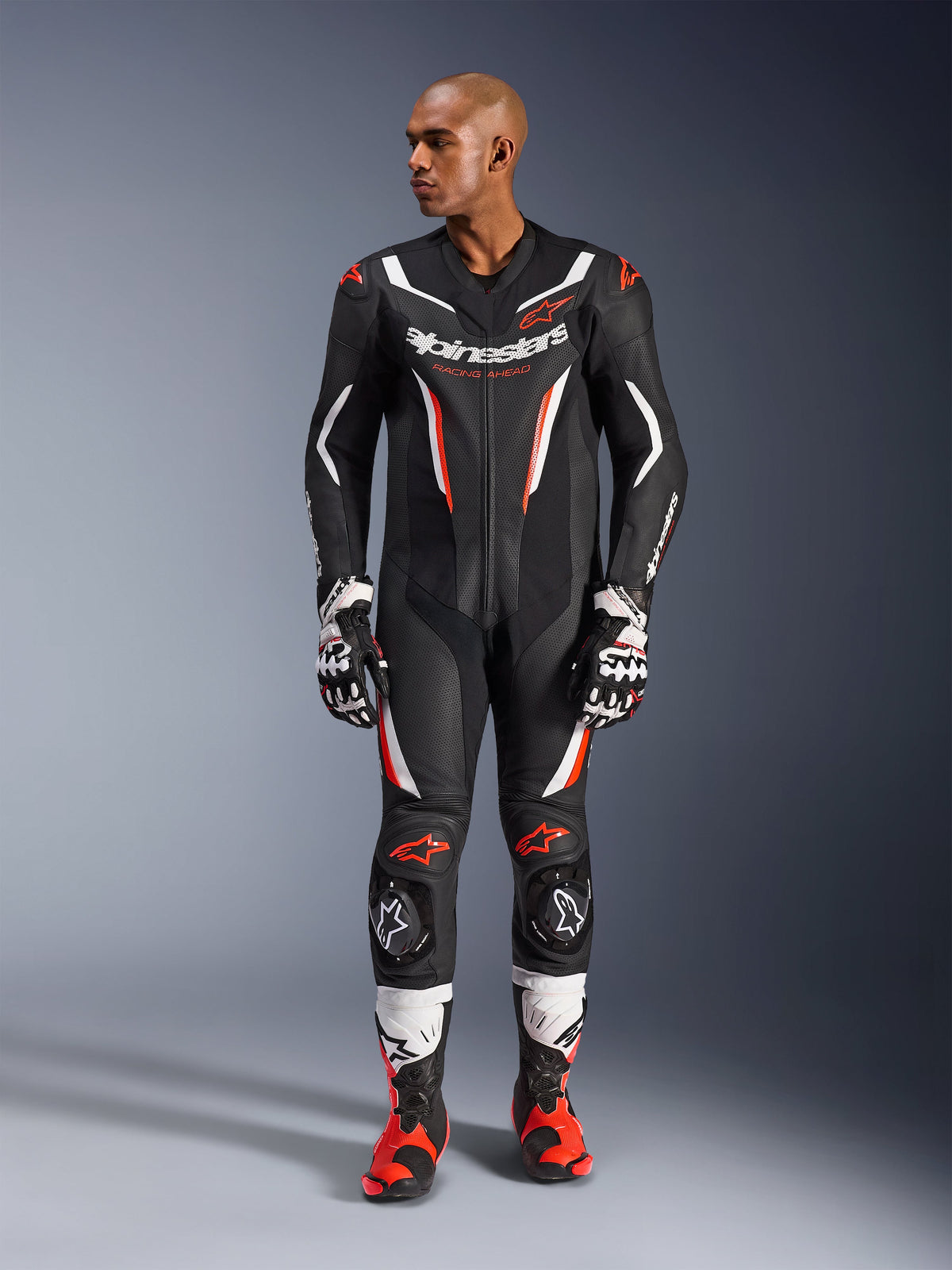 Alpinestars GP Force V2 læderdragt - 1-piece, sort, hvid og rød fluo, helkropsvisning af mandlig model i professionelt motorcykelracerudstyr med matchende handsker og støvler, med perforerede læderpaneler og beskyttende knæslidere.